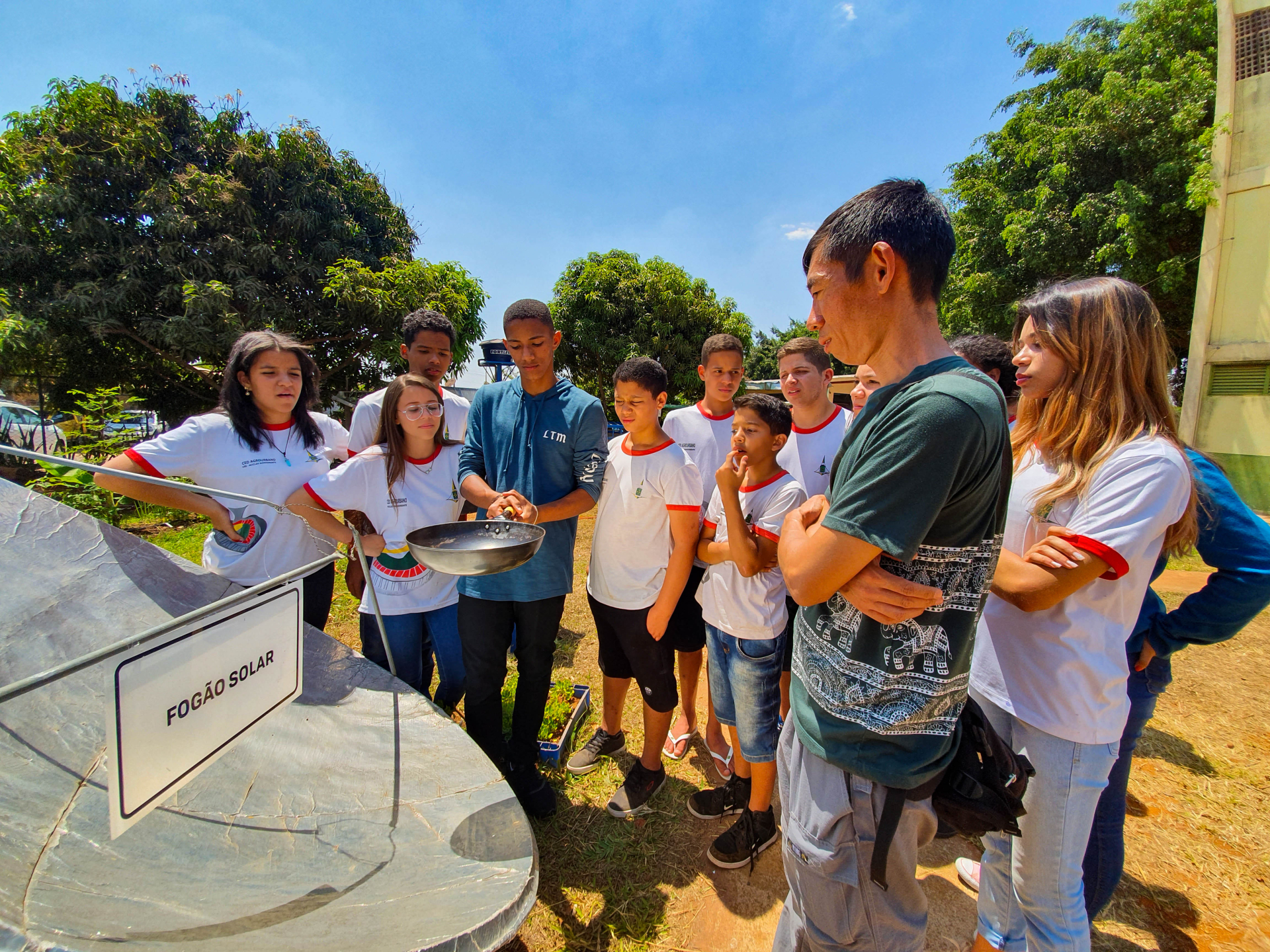 Crianças observam um fogão solar, acompanhadas de um professor.