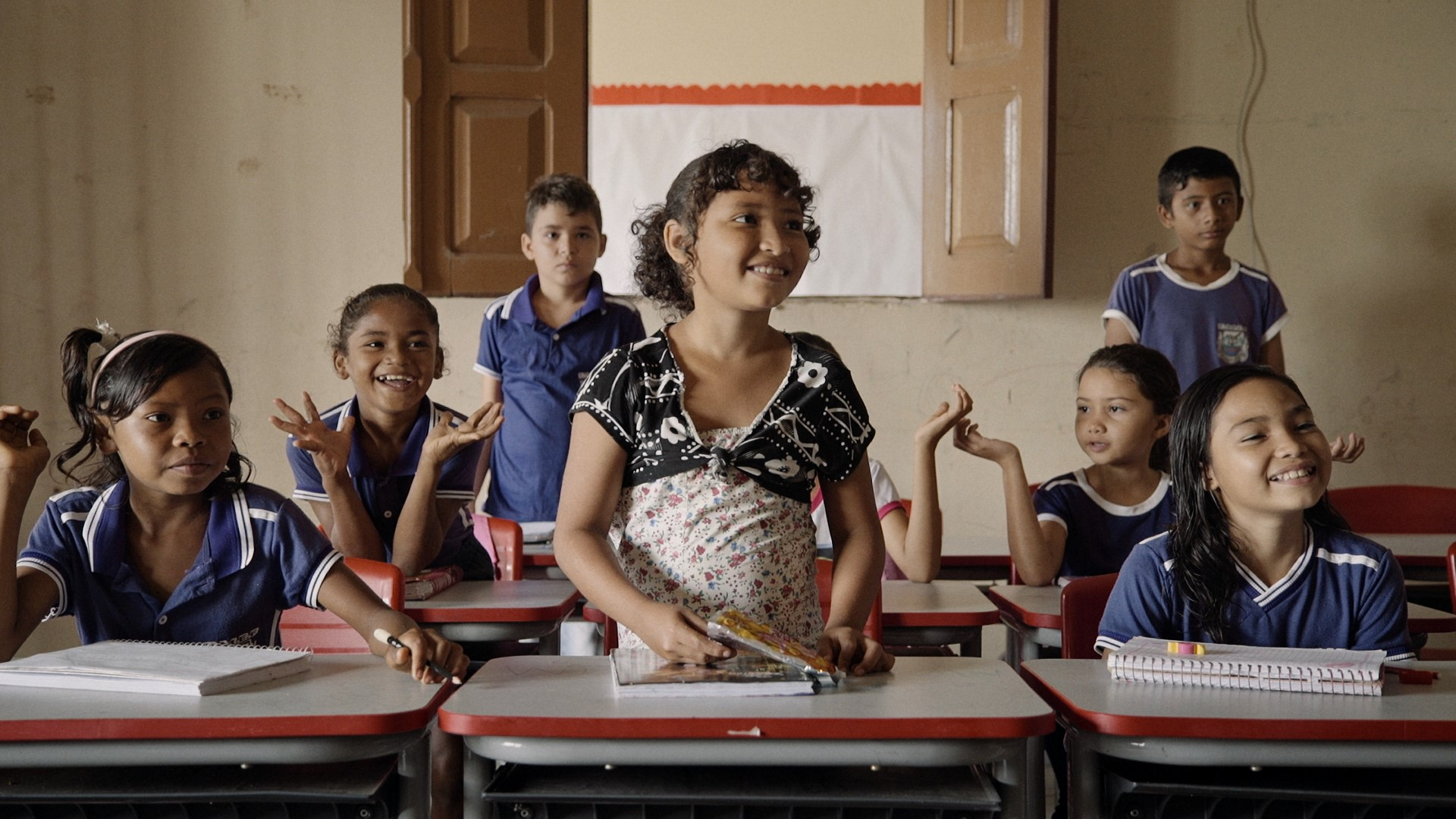 Crianças sorriem em uma sala de aula. Três delas estão de pé.