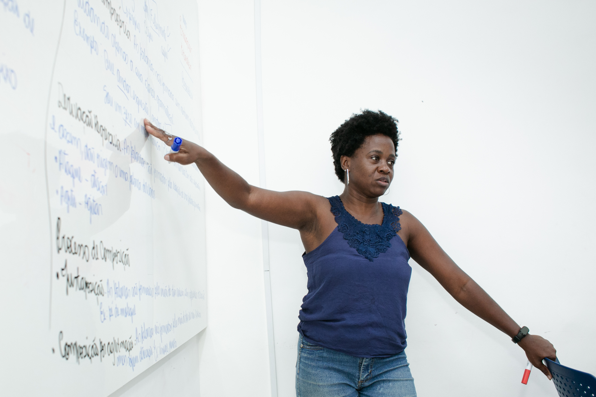 Andrea Ignacio, uma mulher negra, de cabelo curto e preto, com camisa azul e calça jeans, aponta para o quadro em uma sala de aula. Ela está ao lado dele.