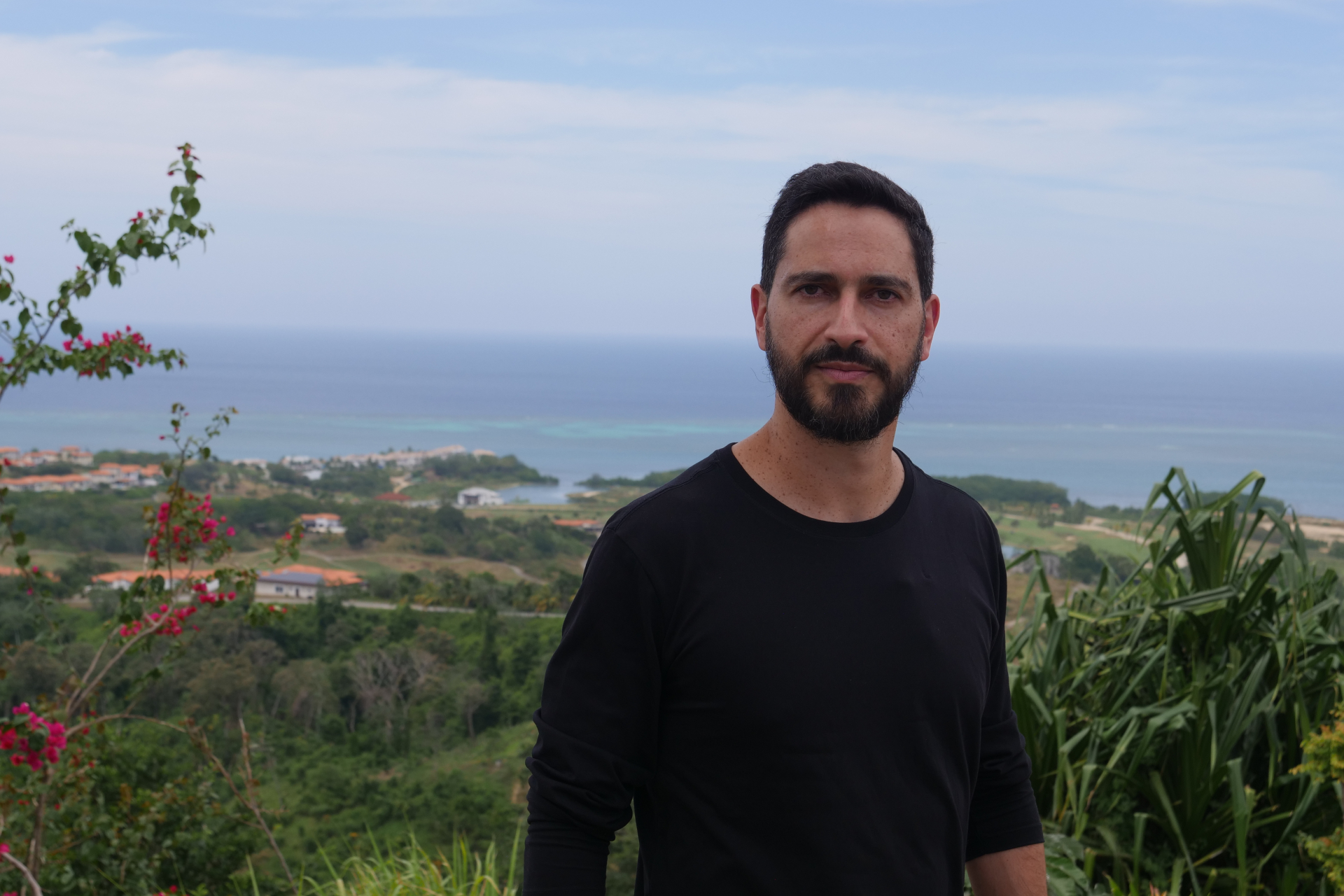 Um homem está de pé. Ao fundo, mais abaixo e distantes, a vegetação e o mar e algumas montanhas.