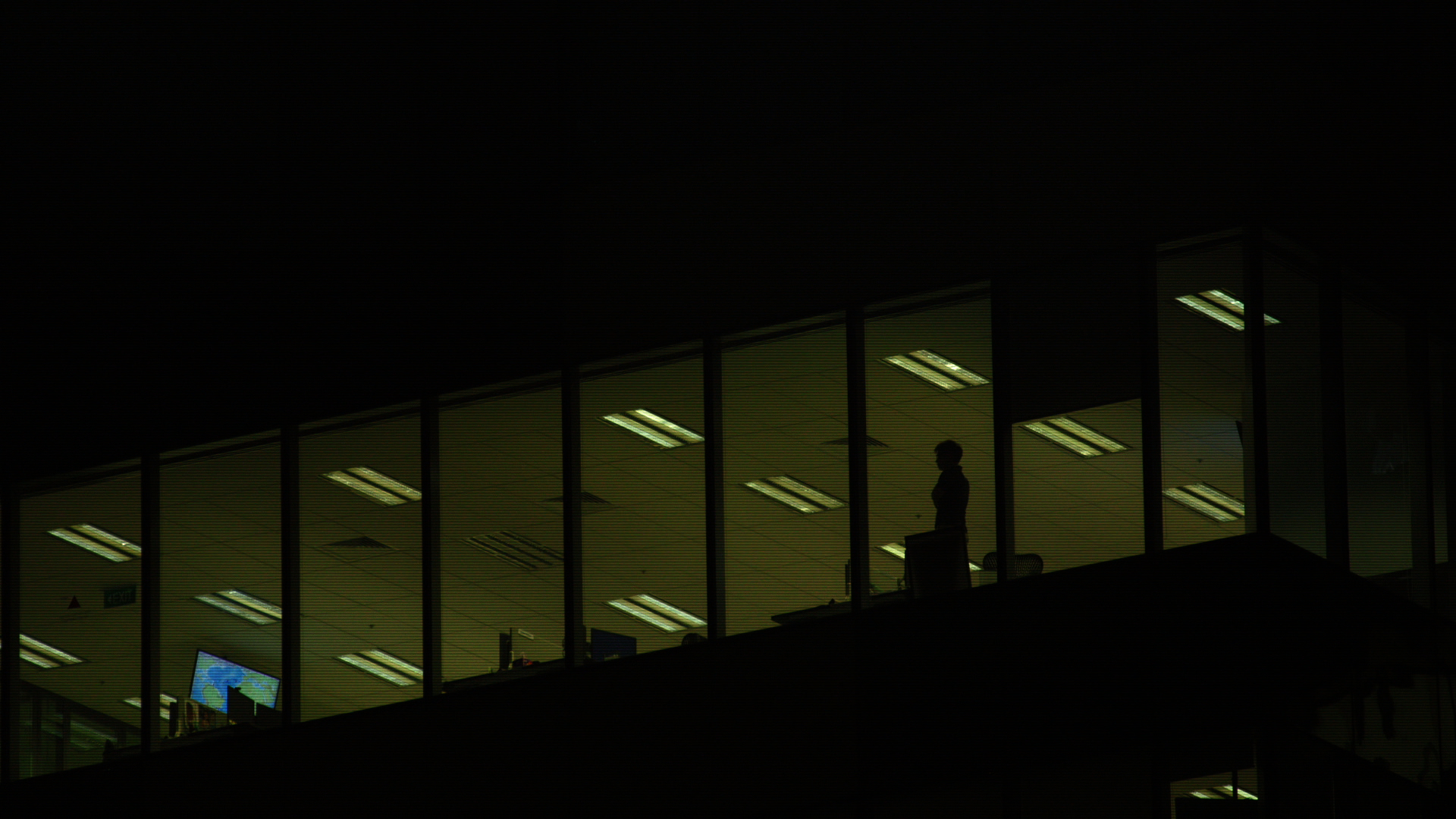 Fotografia noturna de um andar de um prédio comercial. A sala, iluminada, mostra que uma pessoa está lá dentro, de pé.