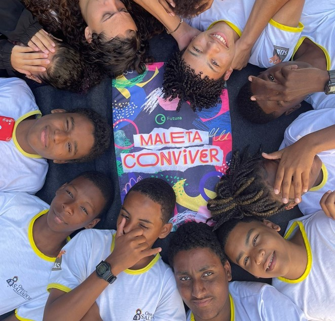 Jovens deitados em círculo (com as cabeças próximas), ao redor de um material colorido com o texto "Maleta Conviver".
