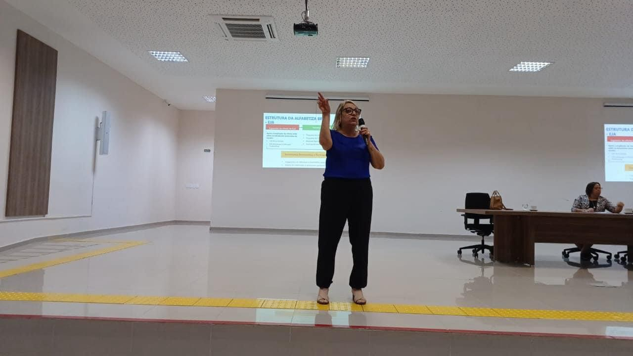 Uma mulher de pé em uma sala clara, com piso de mármore, falando ao microfone.