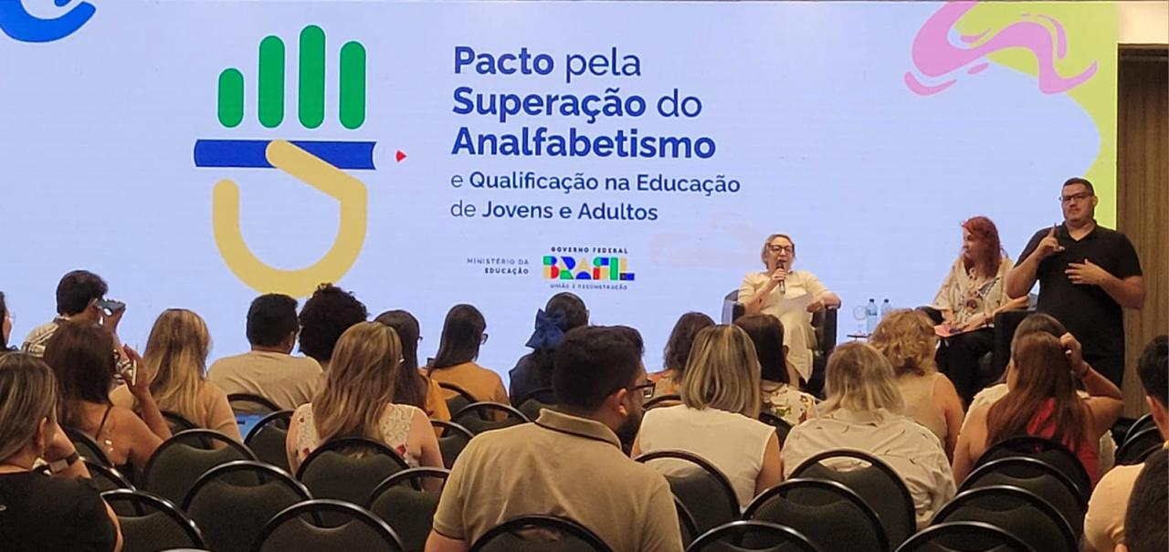Várias pessoas estão na plateia assisindo a uma senhora discursar, sentada.