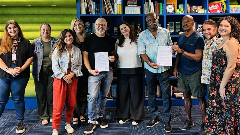 Diversas pessoas reunidas, lado a lado, sorrindo para quem as fotografa. Ao fundo, uma estante com livros.