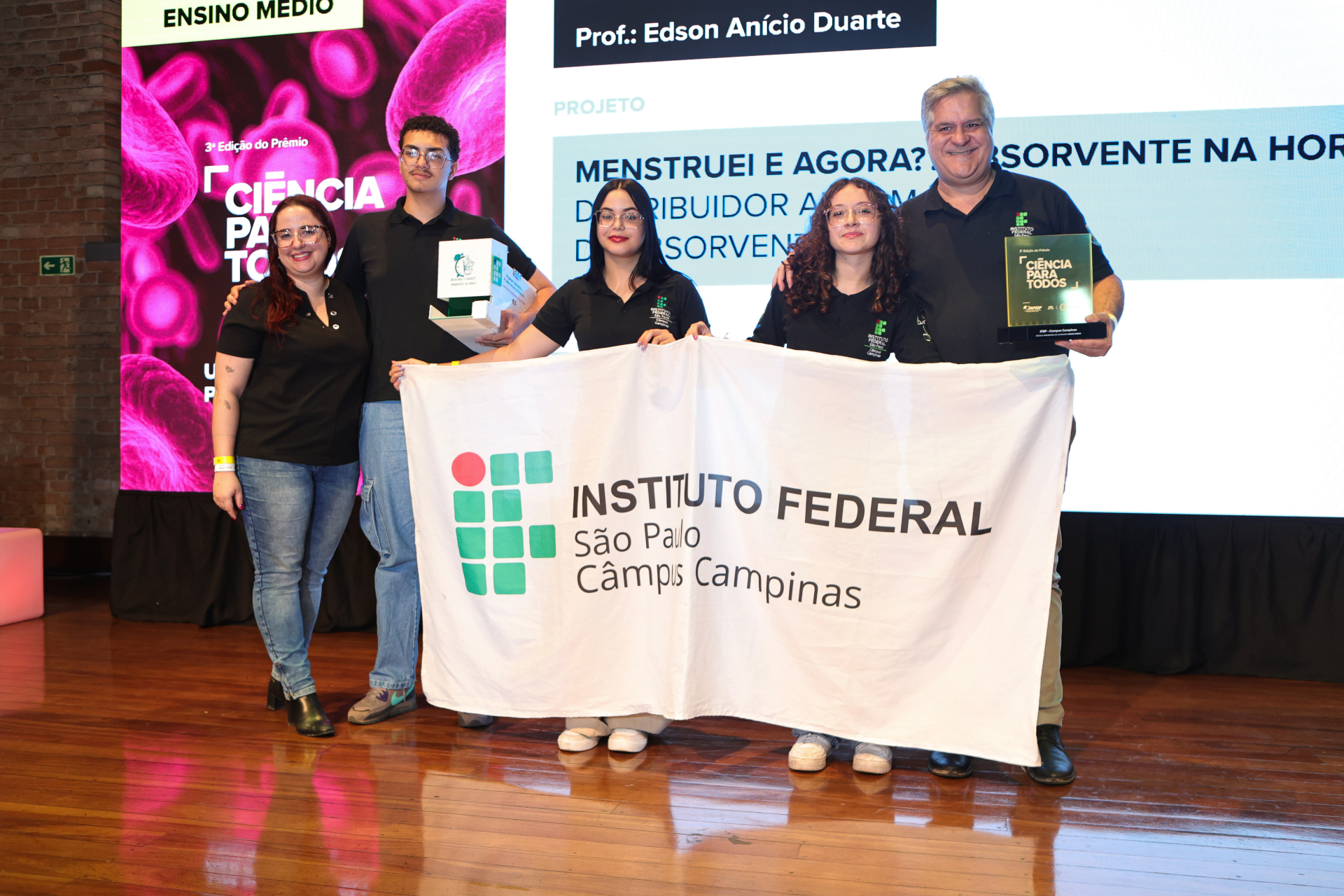 Cinco pessoas de pé em um palco, segurando uma bandeira com o nome do Instituto Federal - Campus Campinas escrito nele.