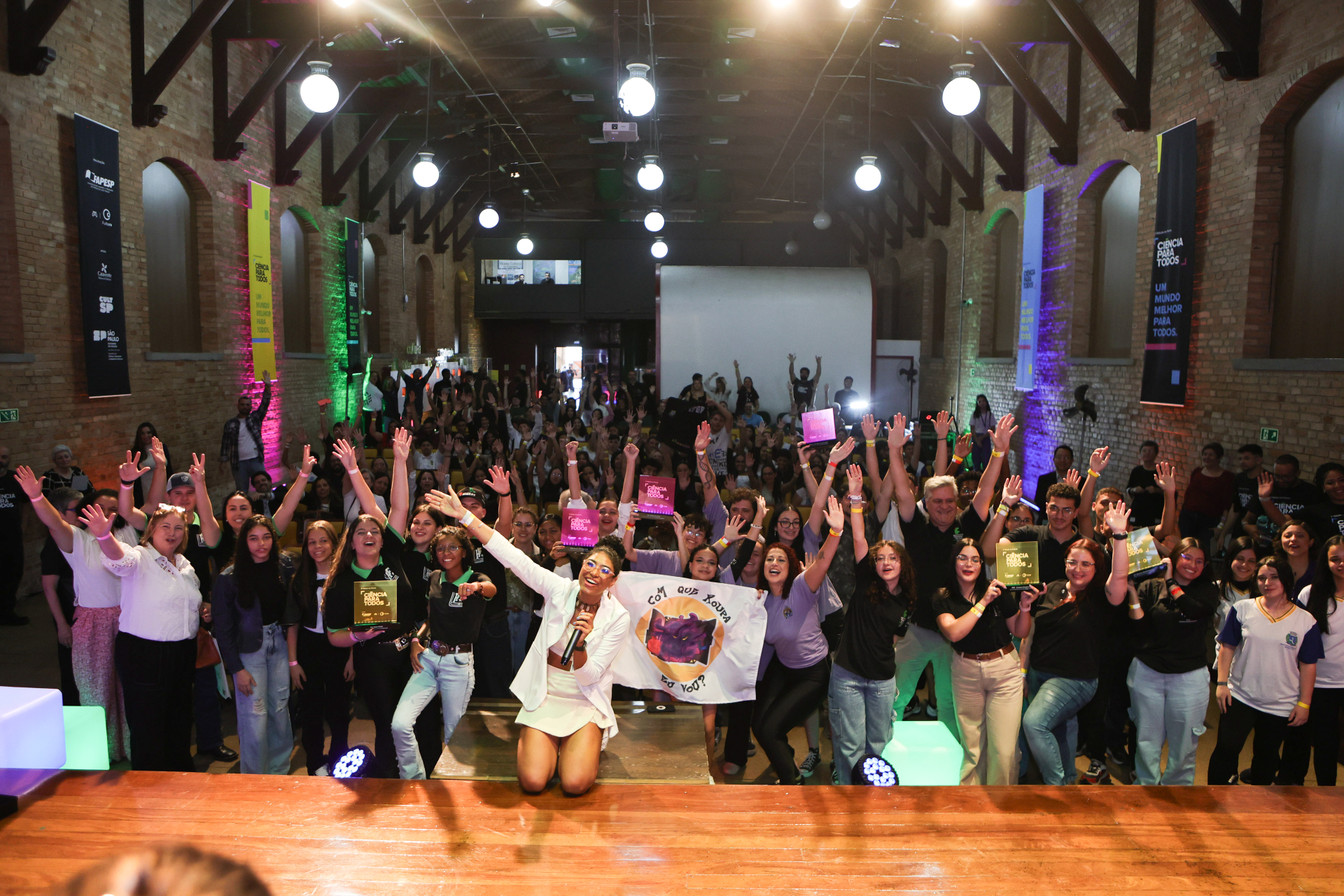 Diversas pessoas com as mãos para o alto, em um auditório. A fotografia foi feita a partir do palco, em direção ao público.