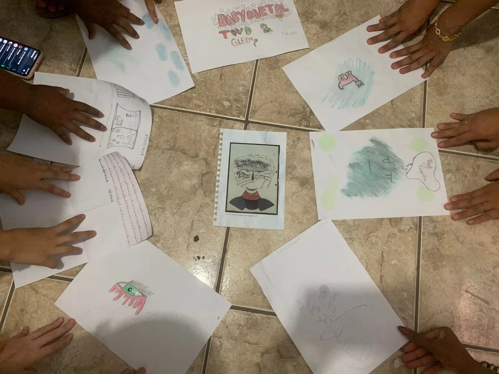 Fotografia captada de cima mostra folhas de papel desenhadas dispostas no chão, com mãos segurando-as.