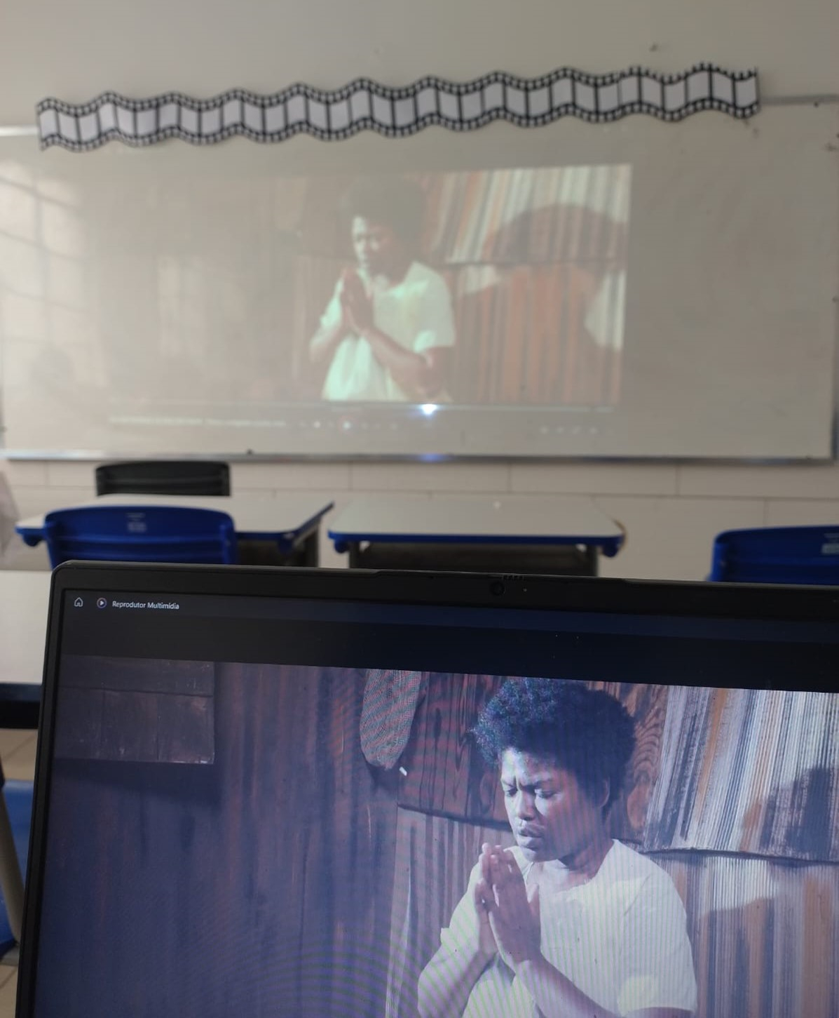 A imagem mostra um notebook no primeiro plano com um vídeo sendo reproduzido e, ao fundo, a mesma imagem sendo projetada em um quadro branco.