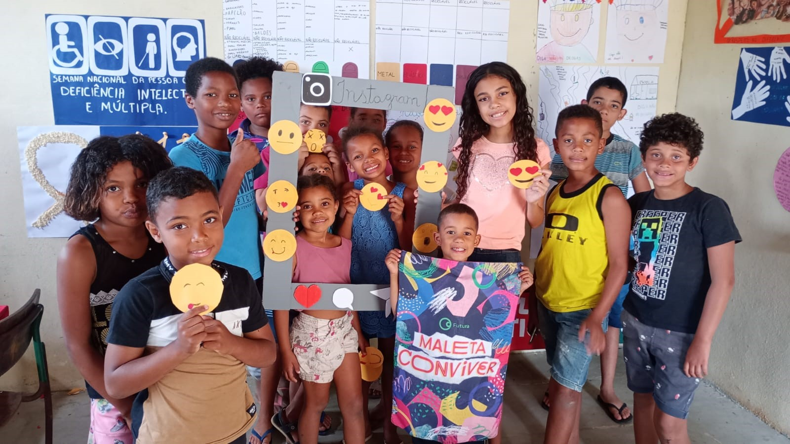 A imagem mostra um grupo de crianças posando e sorrindo para a foto e algumas delas seguram uma moldura com emojis