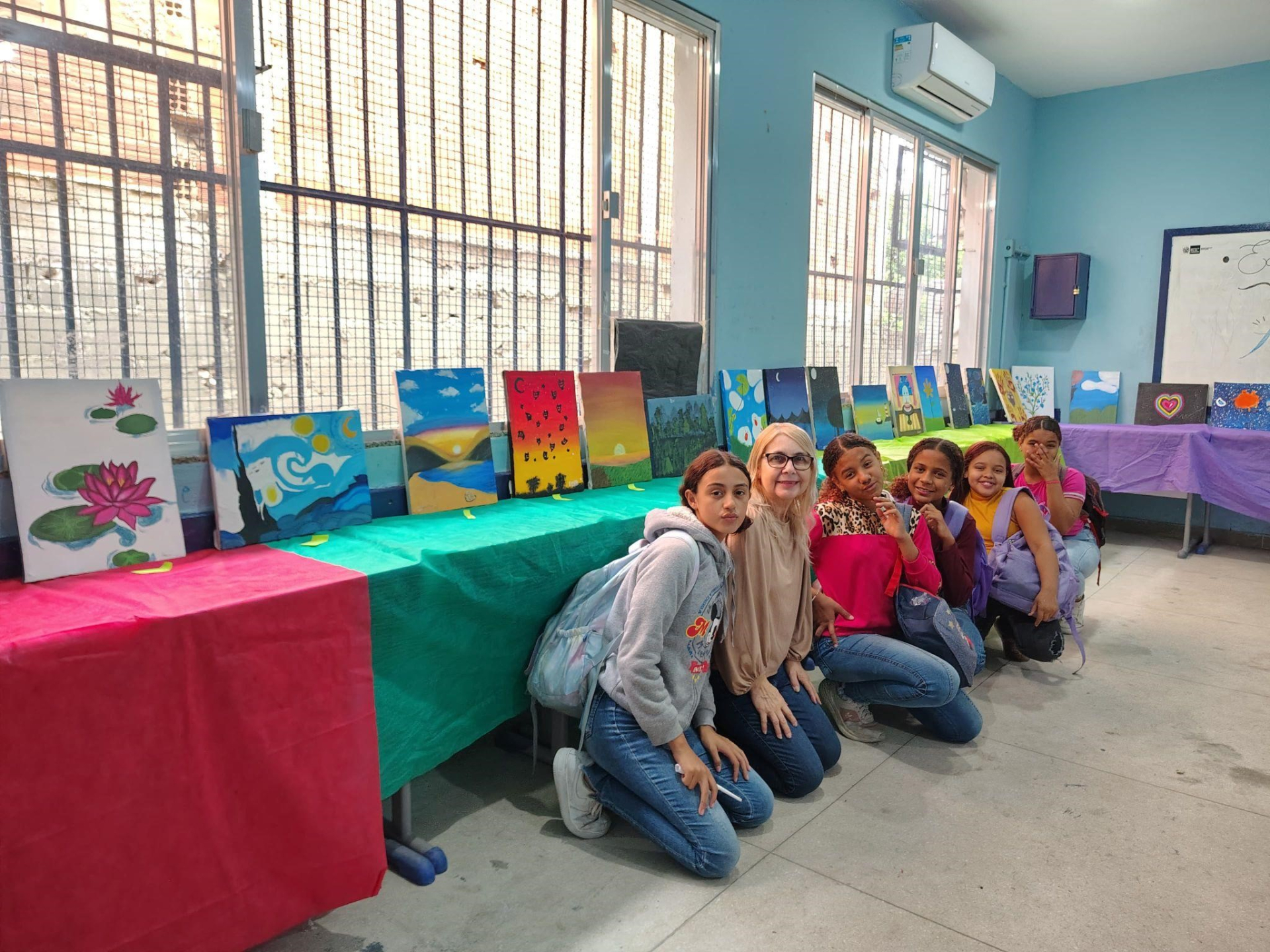 A imagem mostra uma educadora e cinco crianças posando para a foto e agachadas em frente a mesas com pinturas em quadros.