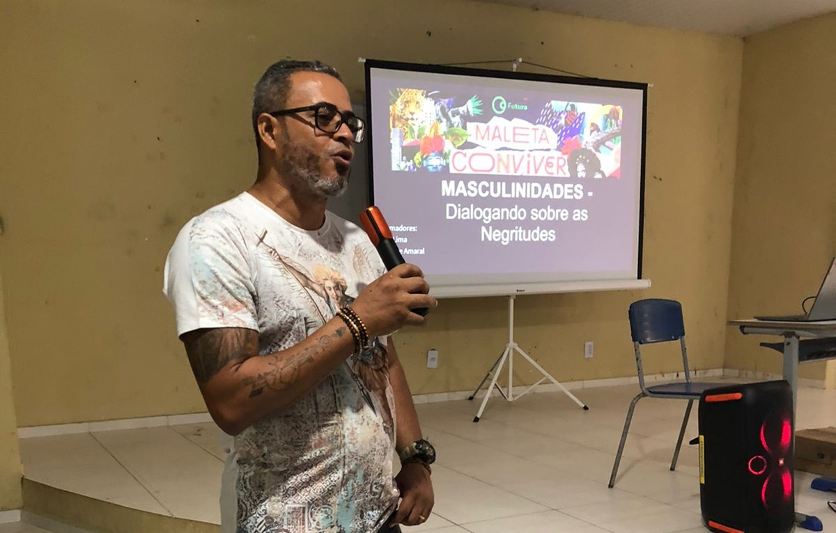 Um homem em uma sala de aula com um microfone na mão. Ao fundo, uma tela retrátil com um slide projetado e com texto escrito "Masculinidades - Dialogando sobre as Negritudes" com a logo do projeto Maleta Conviver.