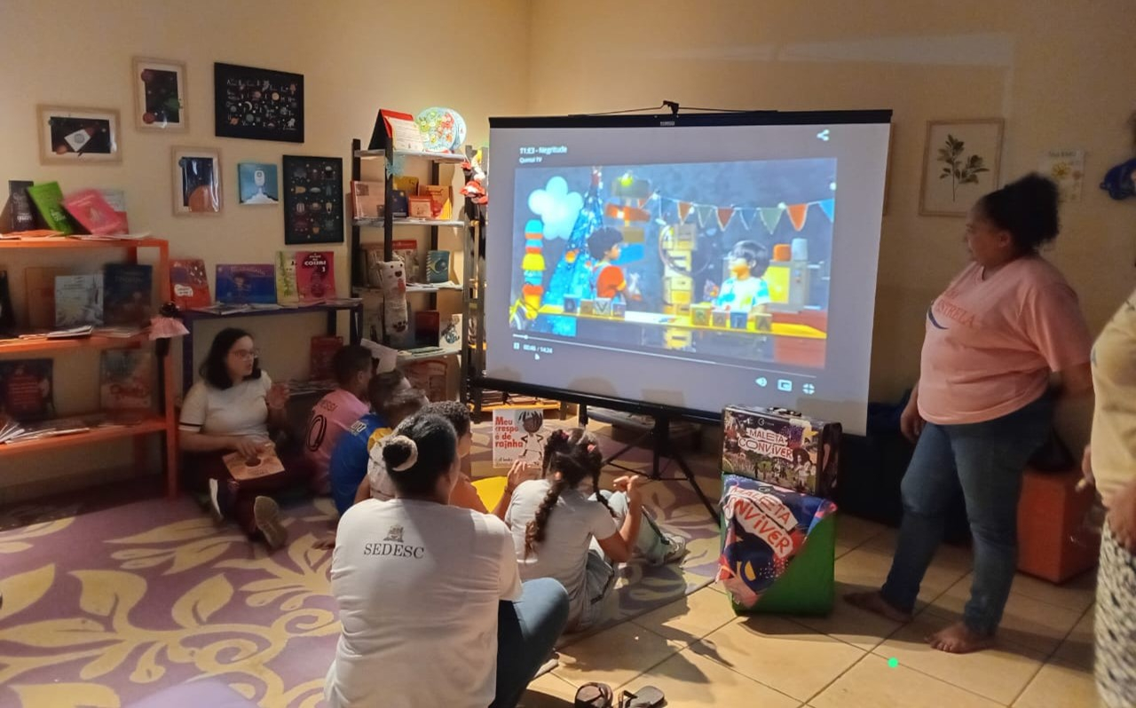 Crianças e educadores em uma sala de aula assistindo um vídeo projetado em uma tela retrátil