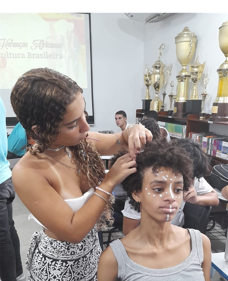 A imagem mostra uma jovem em pé fazendo um penteado no cabelo de outra jovem que está sentada.