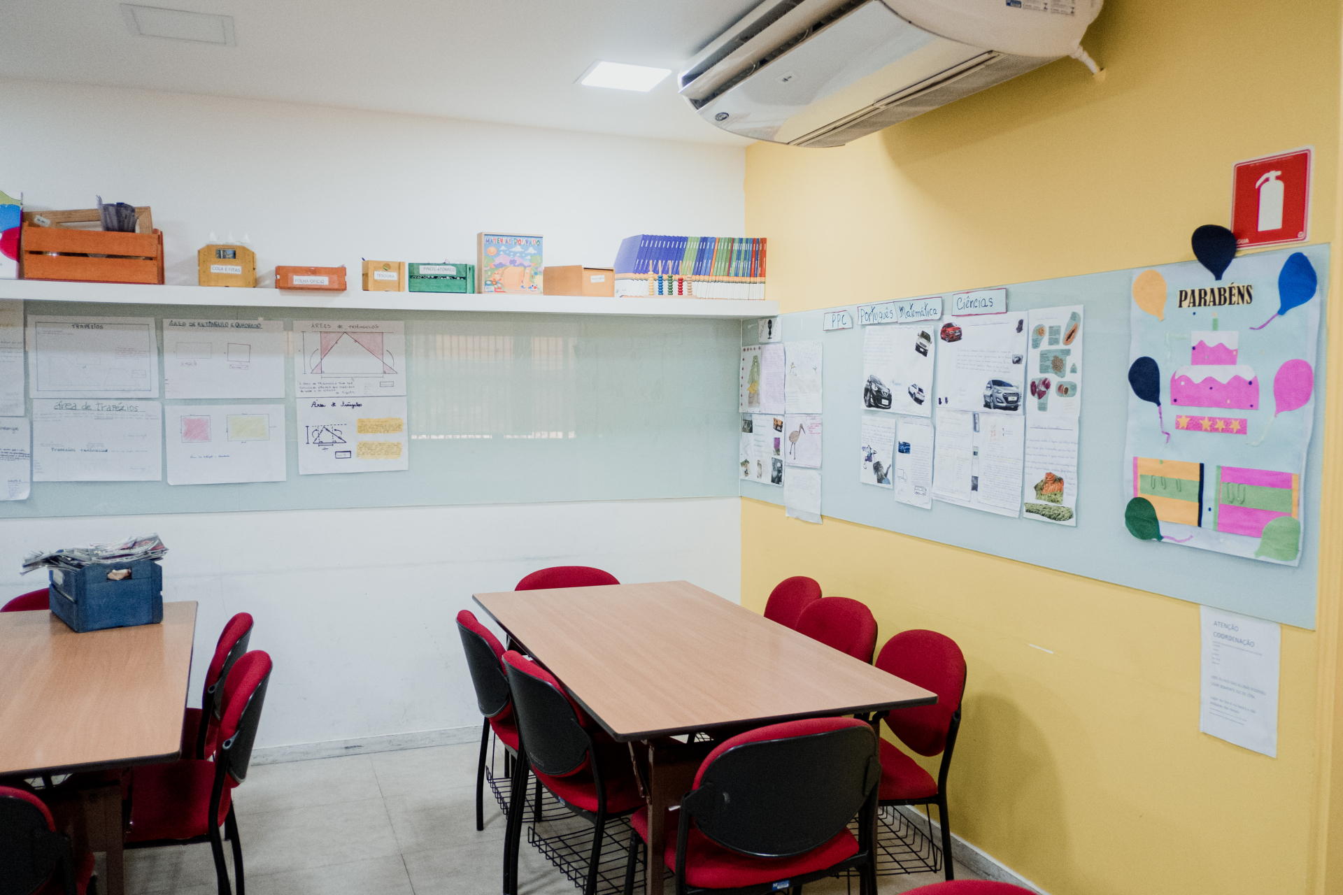 Sala de aula, com mesas e cadeiras. Na parede, há vários cartazes coloridos colados