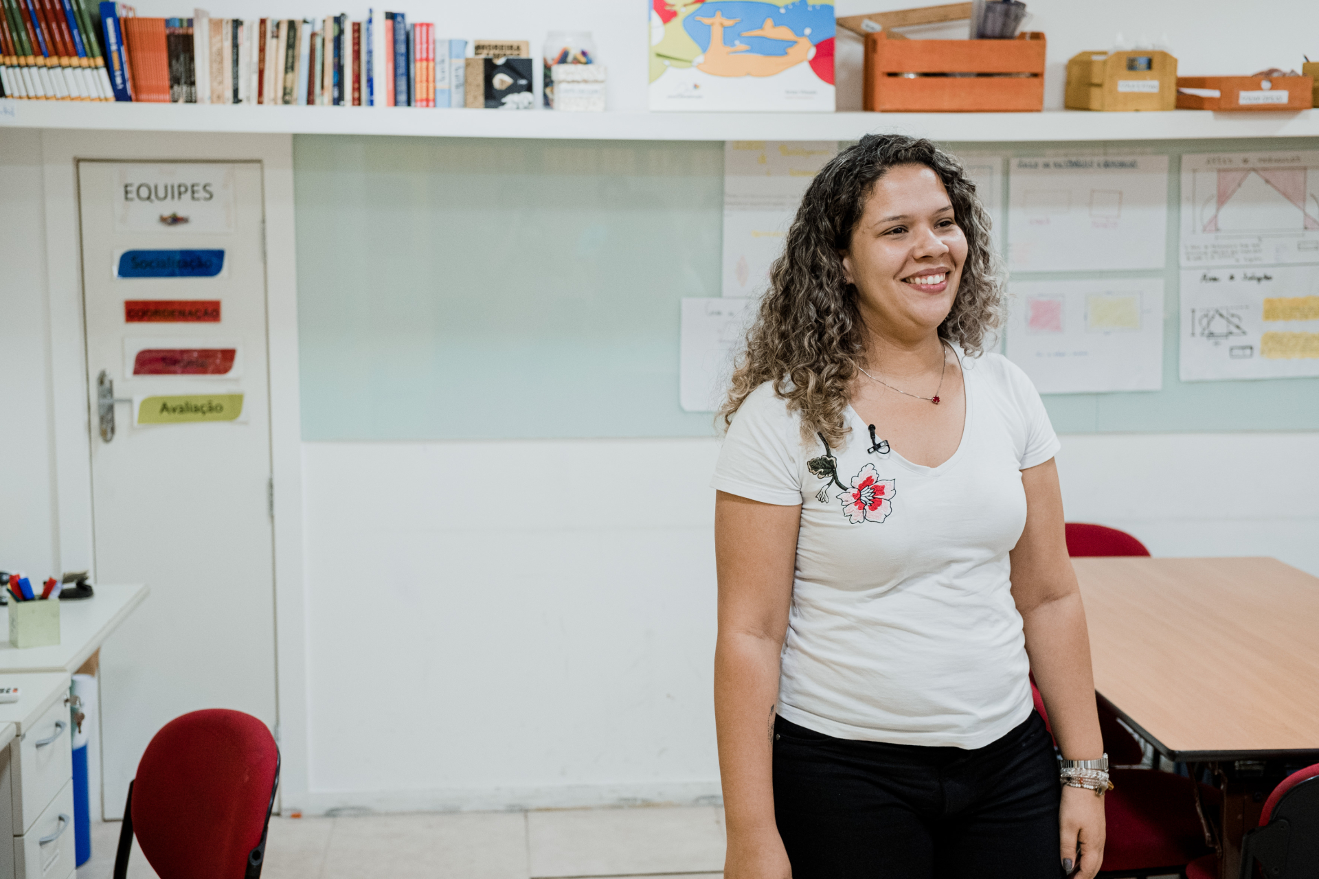 Mulher de cabelos cacheados, blusa branca, em pé no centro de uma sala de aula