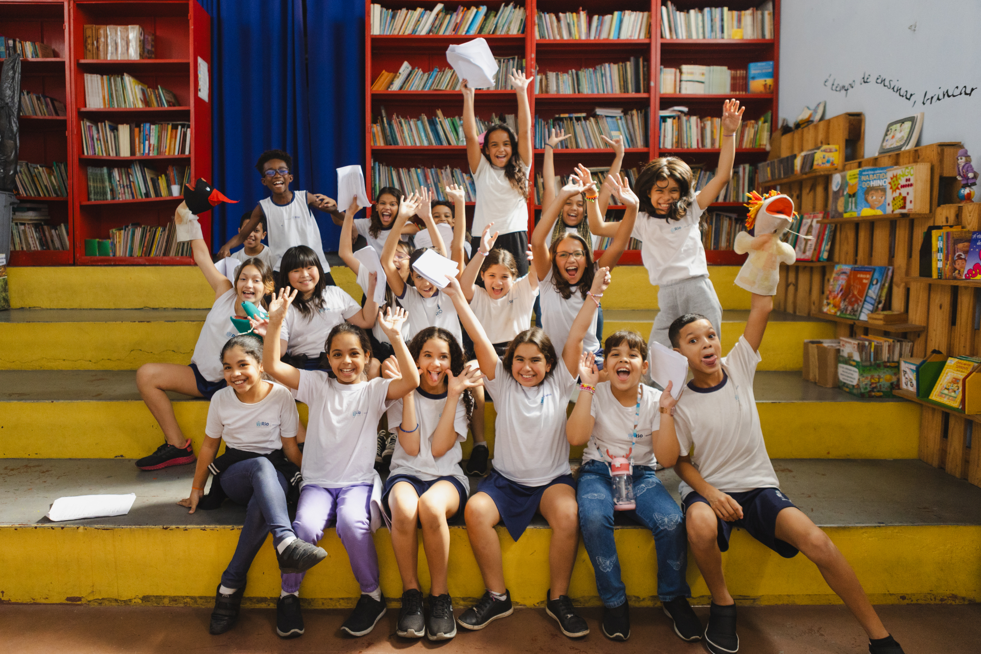 Grupo de estudantes, sentados juntos, com as mãos para cima.