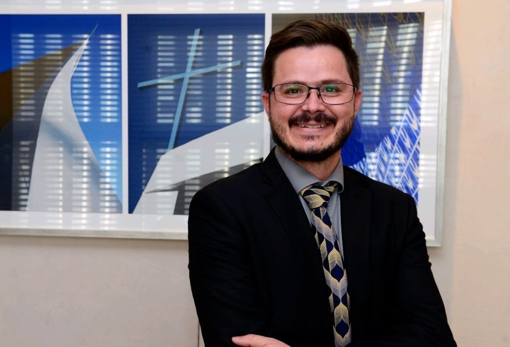 Homem branco, vestindo terno escuro, a frente de um quadro azul
