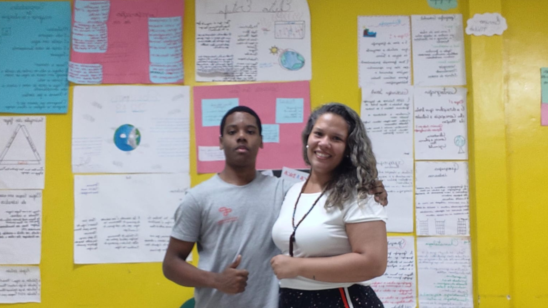 Jovem negro com uniforme escolar e professora de cabelo cacheado a frente de um painel amarelo coberto por cartazes coloridos