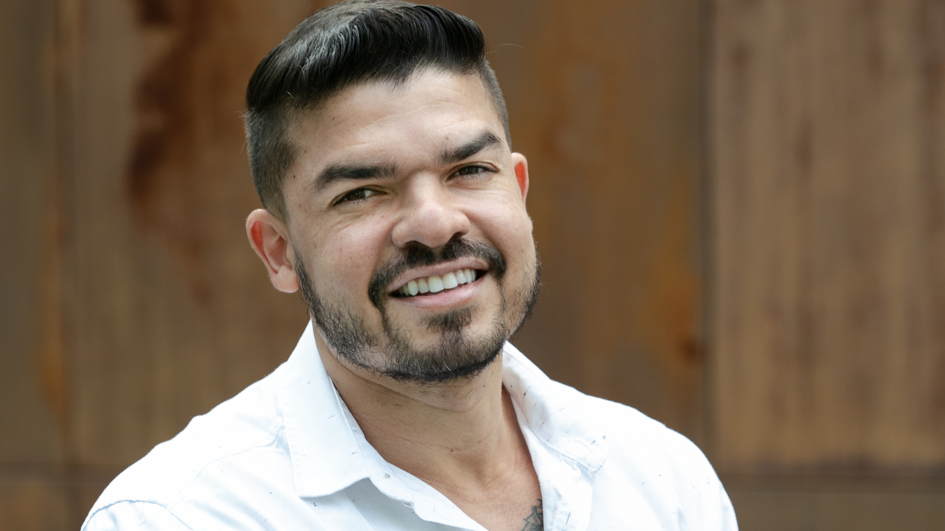 Homem, com barba, vestindo camisa branca a frente de um fundo marrom