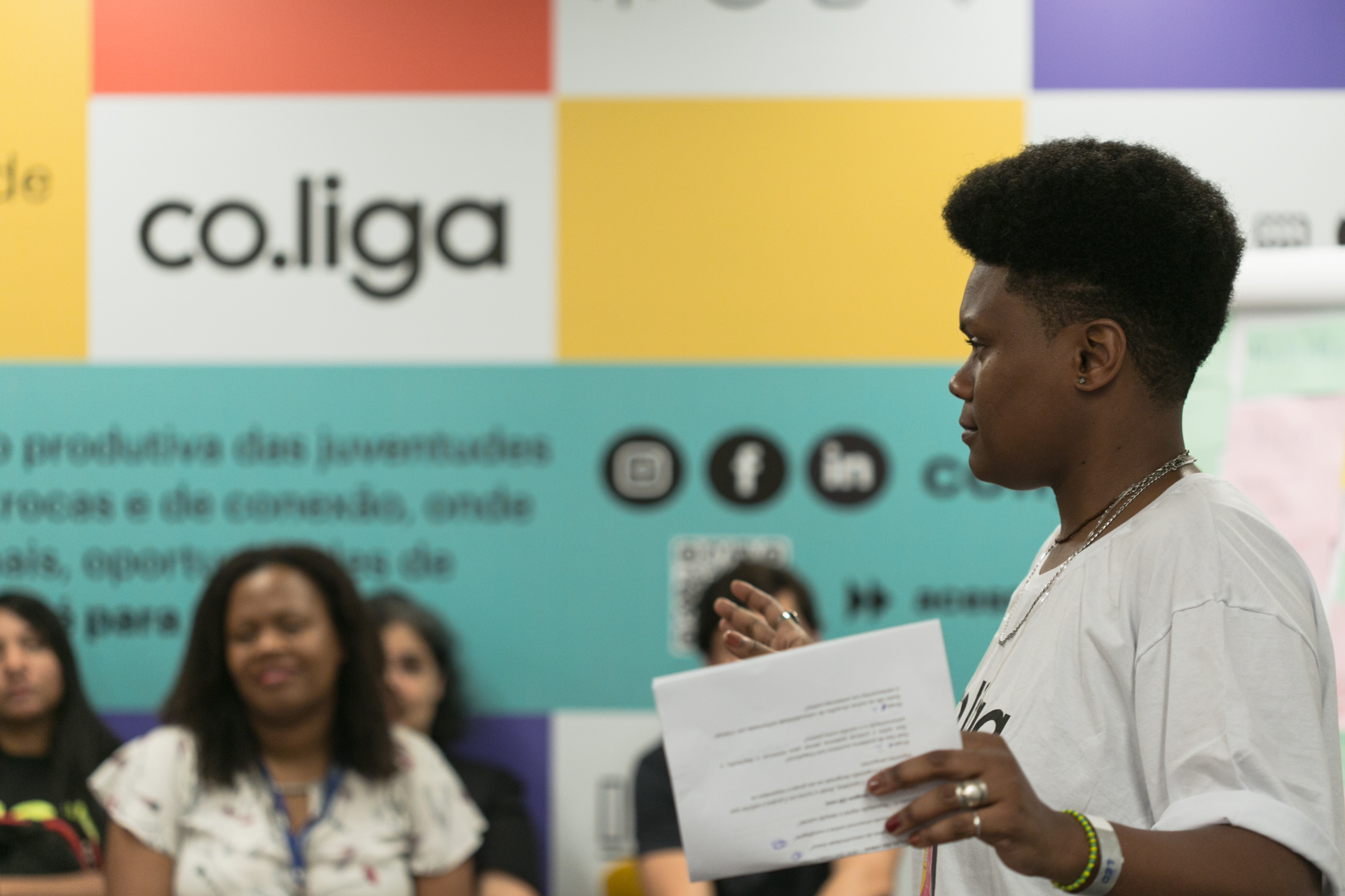 Pessoa negra em pé, de perfil, a frente de um banner colorido da co.liga
