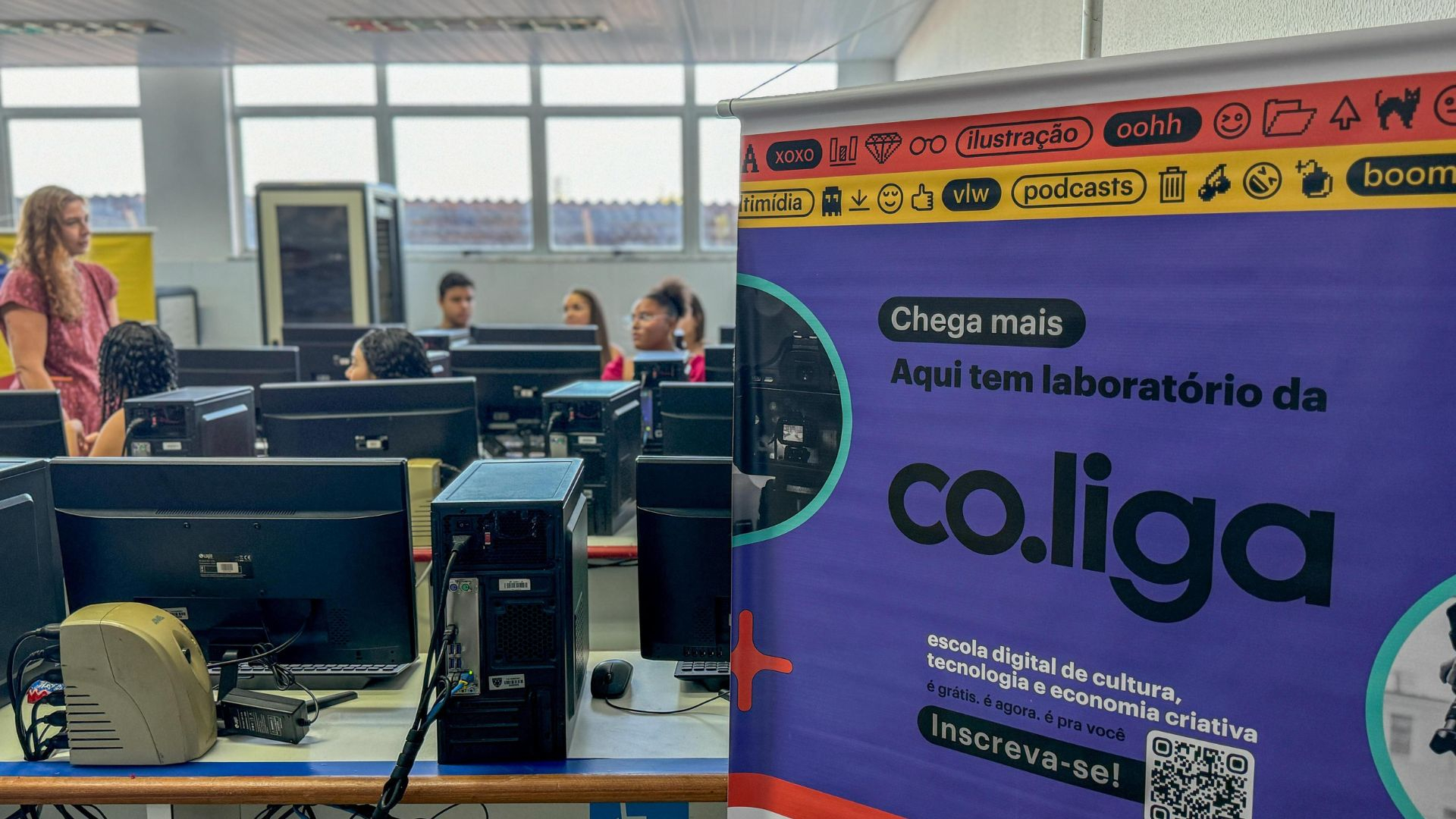 Imagem de um laboratório de informática com jovens participando de uma atividade educativa. No primeiro plano, há um banner colorido da plataforma co.liga, que convida os participantes a se inscreverem em cursos gratuitos de cultura, tecnologia e economia criativa. Ao fundo, uma sala com vários computadores e jovens atentos, enquanto uma facilitadora, em pé, interage com o grupo. A luz natural entra pelas janelas amplas, iluminando o ambiente de aprendizado e troca.