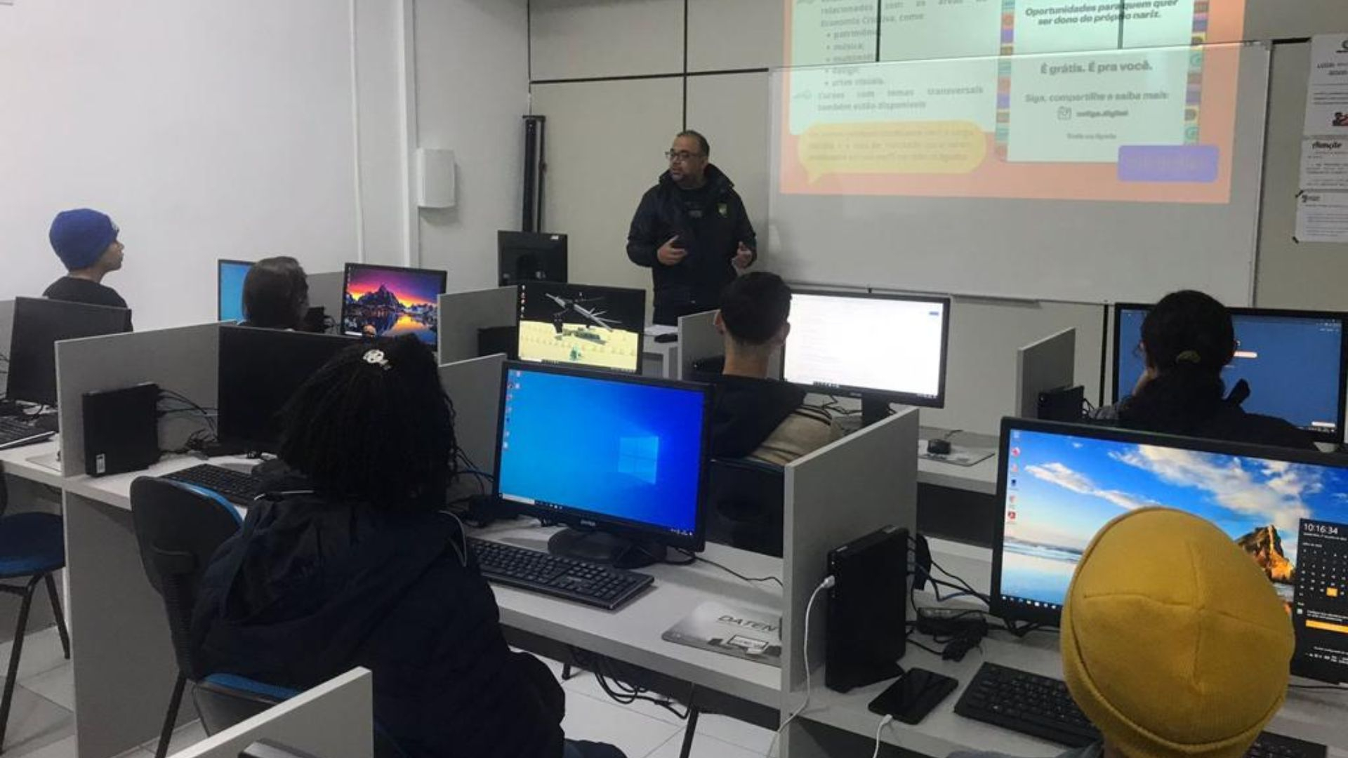 Turma de estudantes participa de aula em laboratório de informática, com foco em tecnologia e inclusão digital. A imagem mostra um educador à frente da sala, explicando um conteúdo projetado na parede, enquanto os alunos acompanham atentamente de suas estações de trabalho, cada uma equipada com computador. O ambiente é moderno, com divisórias individuais que favorecem a concentração. A aula faz parte de uma iniciativa educacional gratuita, voltada para formação técnica e desenvolvimento de competências digi