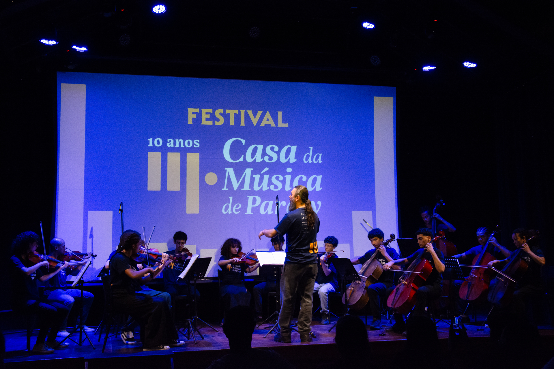 No palco de um teatro, um grupo de jovens músicos se apresenta sob a regência de um maestro. Eles tocam instrumentos de corda, como violinos, violoncelos e contrabaixos. Todos estão usando camisetas pretas com a identidade visual do evento. Ao fundo, um grande telão projeta o nome do festival: "Festival 10 anos Casa da Música de Paraty", com fundo azul e letras em branco e amarelo. A iluminação azul suave cria uma atmosfera intimista e acolhedora, e há público assistindo na plateia, visível em silhueta na p