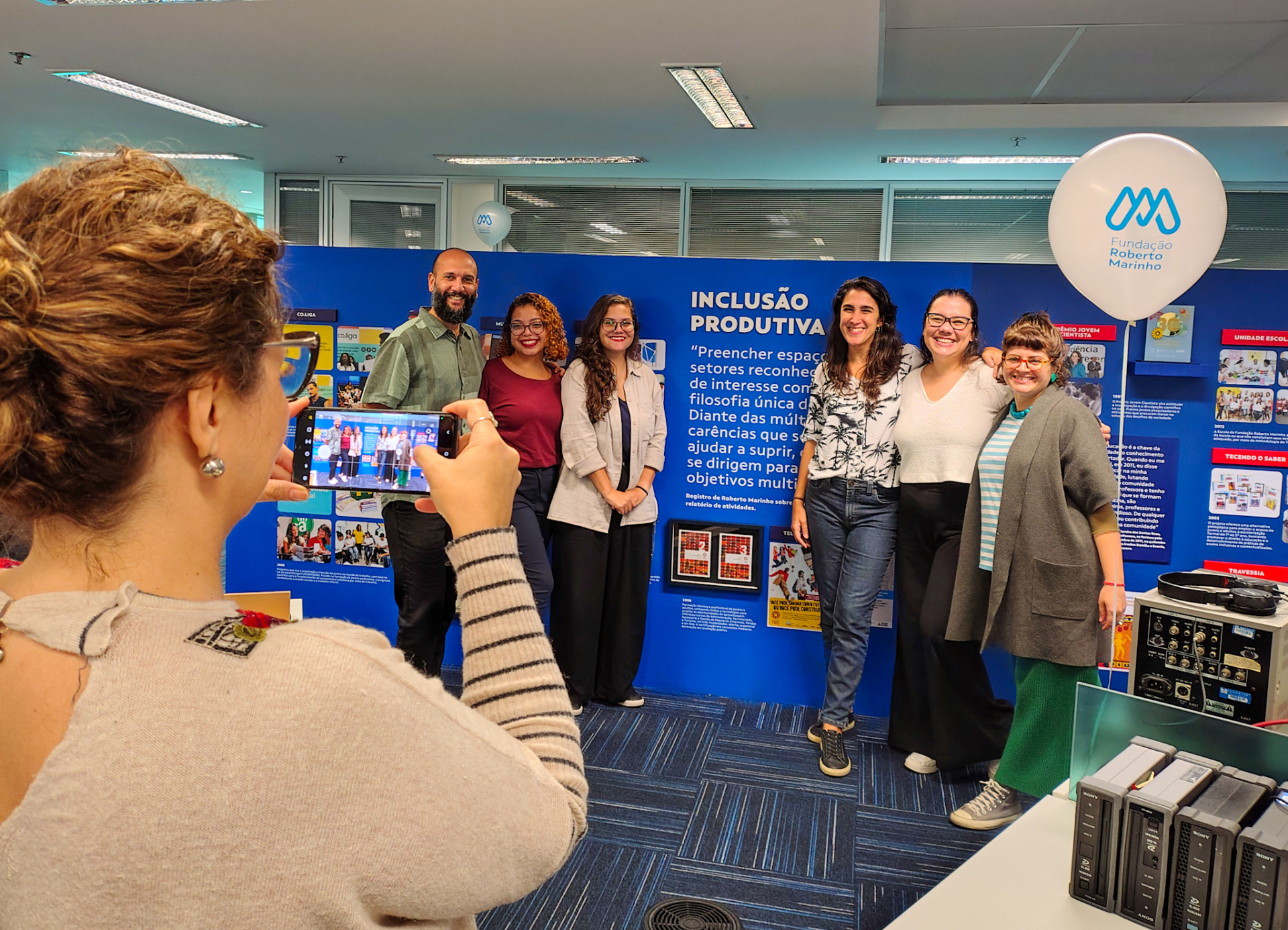 Funcionária fotografa um grupo de seis colegas sorridentes em frente a um painel com o tema “INCLUSÃO PRODUTIVA”, na sede da Fundação Roberto Marinho. O painel contém textos, imagens e um balão decorativo com o logotipo da instituição. O ambiente expressa celebração, pertencimento e valorização do trabalho em equipe
