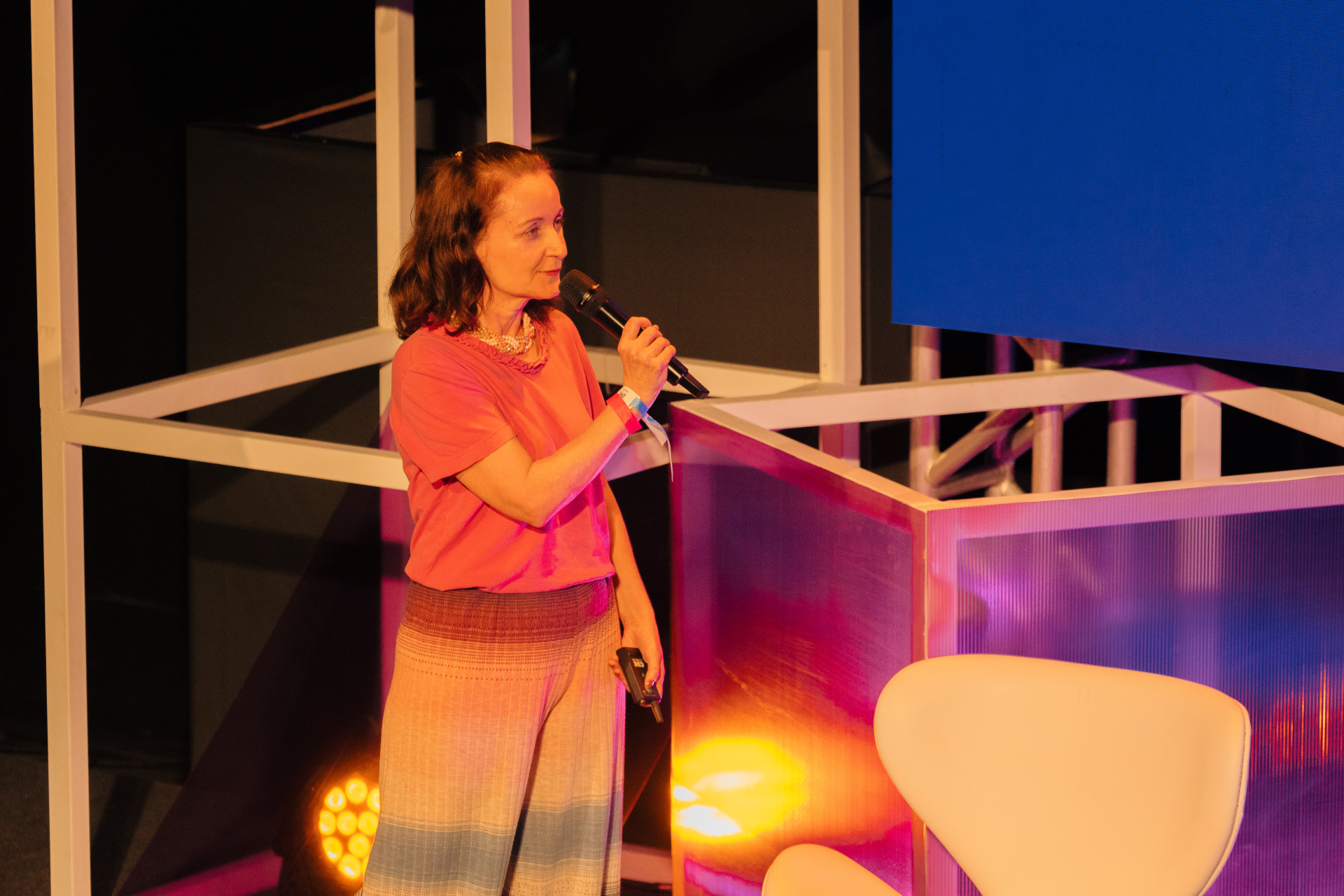 Rosalina Soares, superintendente de Conhecimento da Fundação Roberto Marinho, participa como palestrante em um evento. Ela segura um microfone na mão e está a frente de uma estrutura metálica branca que parece ser um palco.
