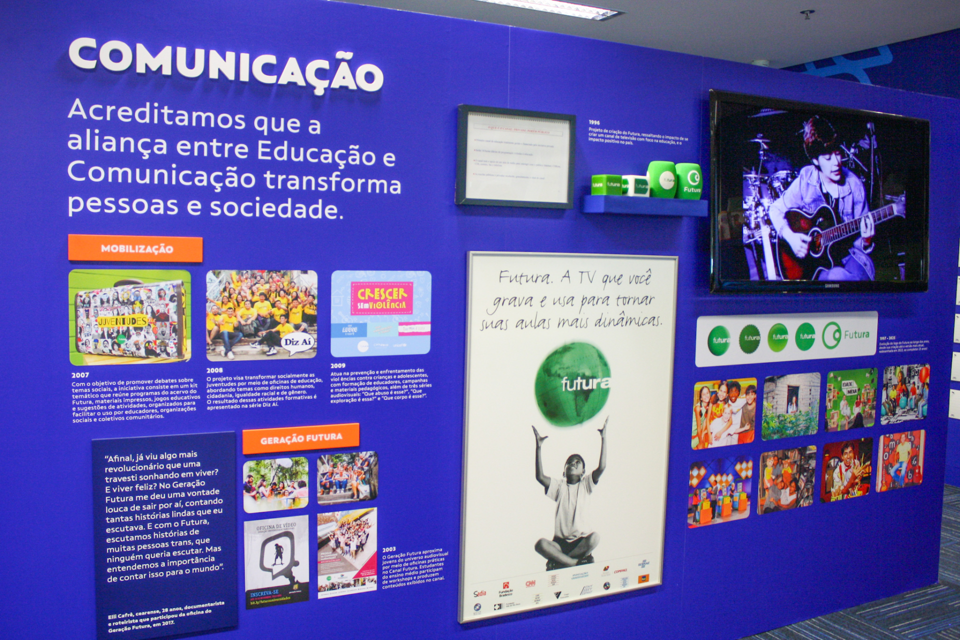 Painel expositivo sobre Comunicação e Educação, com destaque para o Canal Futura, em um ambiente interno iluminado. O painel, de cor azul vibrante, apresenta textos, imagens, vídeos e cartazes que mostram a importância da comunicação como ferramenta de transformação social. No topo, lê-se a frase: “Acreditamos que a aliança entre Educação e Comunicação transforma pessoas e sociedade.”  À esquerda, há seções intituladas "Mobilização" e "Geração Futura", com fotos de projetos educativos e sociais realizados e