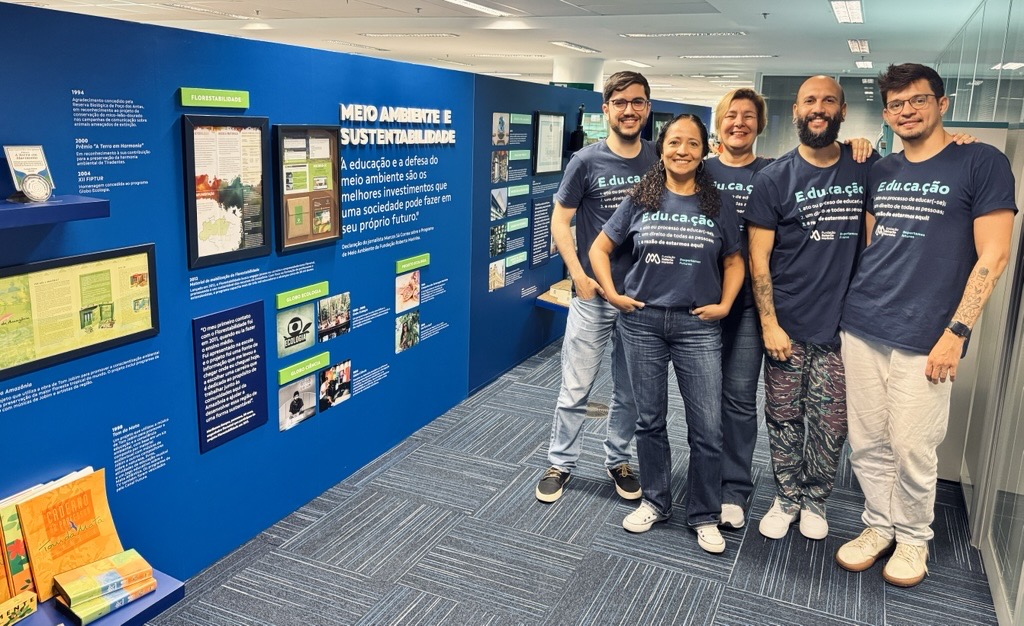 Cinco colaboradores posam sorridentes em frente a um painel temático com o título “MEIO AMBIENTE E SUSTENTABILIDADE”. Todos vestem camisetas com a definição da palavra “Educação”, reforçando o compromisso da Fundação Roberto Marinho com a conscientização ambiental e a formação cidadã. O painel exibe projetos, publicações e ações educativas ligadas à causa.