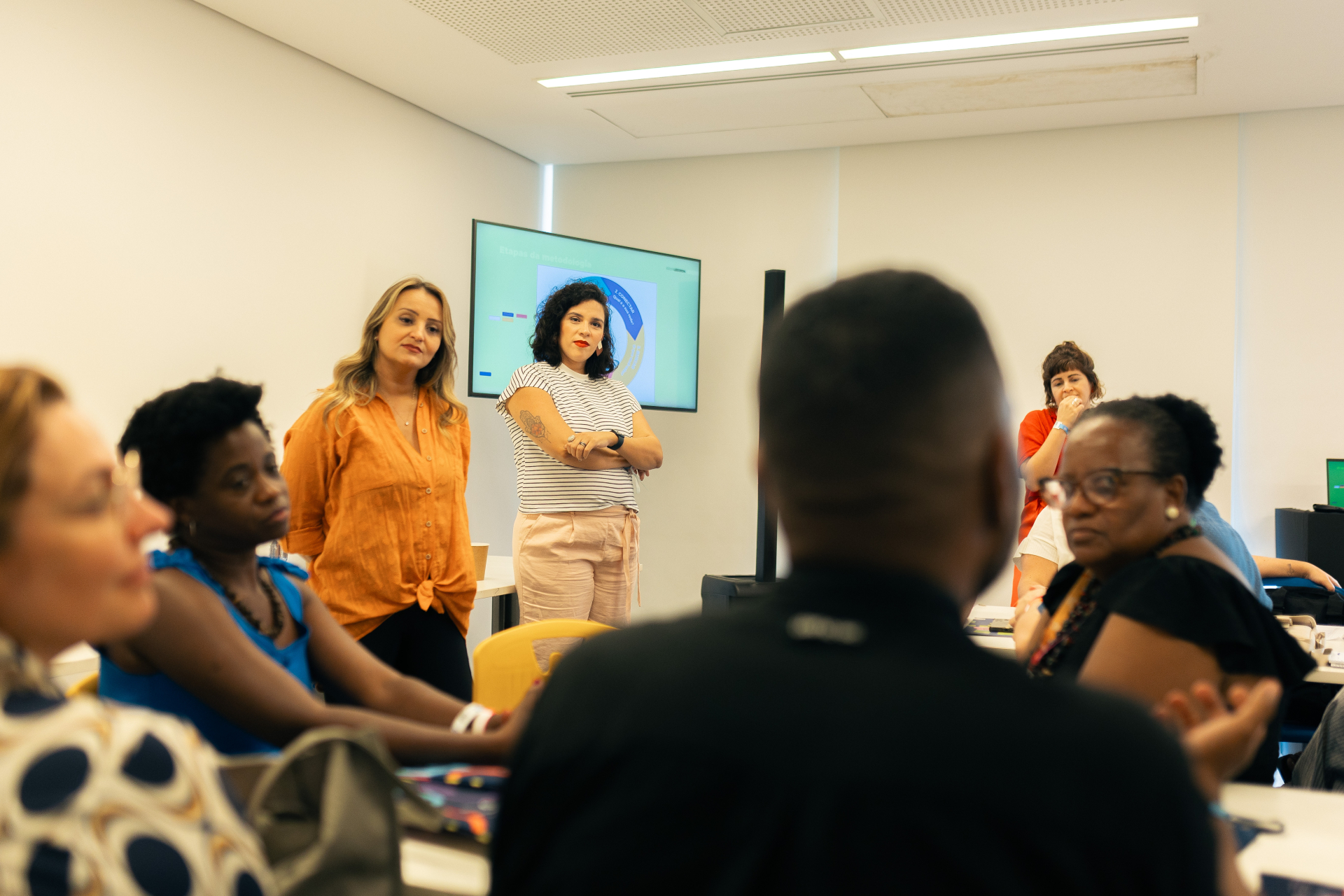 Em uma sala de reunião bem iluminada, um grupo diverso de pessoas está reunido em um ambiente colaborativo. Duas mulheres em pé, ao fundo, observam atentamente e interagem com os participantes sentados, enquanto uma tela exibe uma apresentação com gráficos e textos parcialmente legíveis. Uma terceira mulher, também em pé, parece refletir ou escutar com atenção, com a mão no queixo. As demais pessoas, sentadas ao redor de uma mesa, participam da conversa com expressões de interesse e engajamento. O ambiente