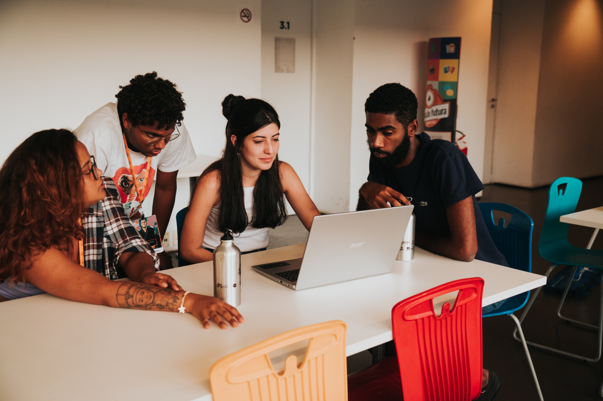 A imagem mostra quatro jovens conversando e olhando algo em um notebook