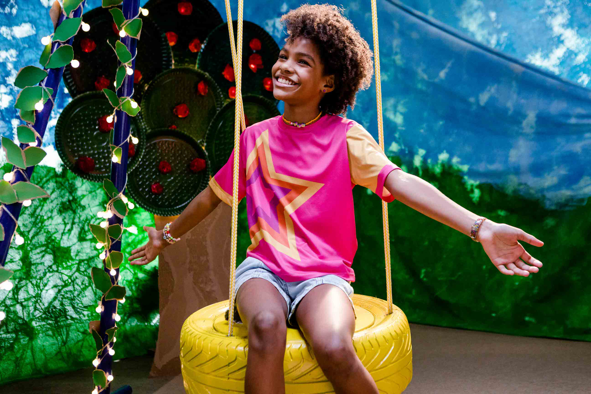 Menina negra sorridente, de cabelos cacheados curtos, sentada em um balanço feito de pneu amarelo pendurado por cordas. Ela usa camiseta rosa com estampa de estrela amarela e mangas curtas em tom alaranjado, além de short jeans. Ao fundo, um cenário colorido com pinturas que lembram céu, árvores e folhagens, além de um tronco decorado com folhas verdes artificiais e pequenas luzes.