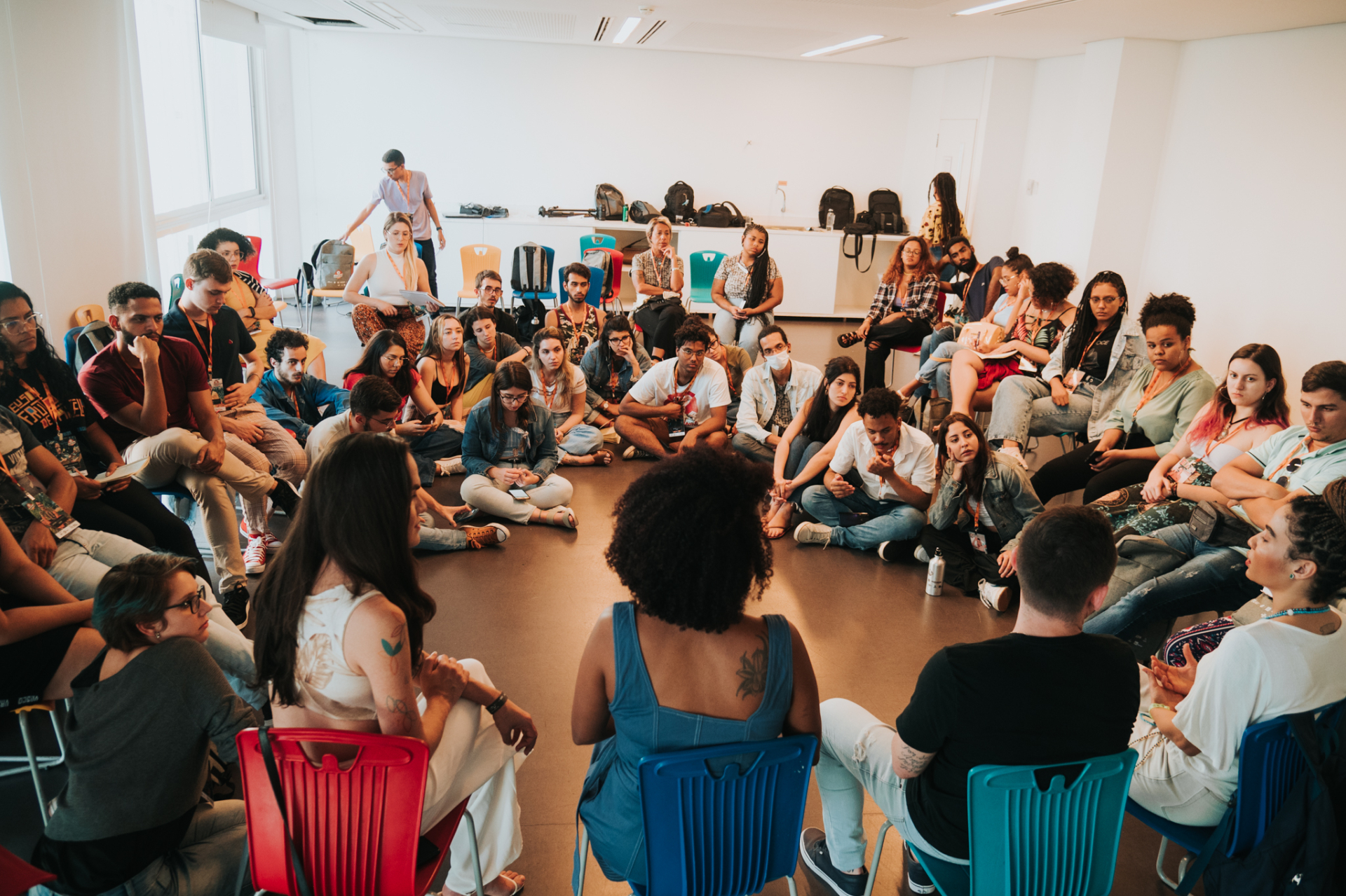 Sala ampla e iluminada, com cerca de 40 jovens sentados em círculo no chão ou em cadeiras coloridas. Todos estão voltados para o centro da roda, participando de uma conversa coletiva. As expressões variam entre atenção, curiosidade e escuta ativa. Ao fundo, há mochilas e objetos pessoais sobre uma bancada branca. A cena transmite um ambiente acolhedor, de partilha e escuta, típico de uma atividade formativa ou roda de conversa.