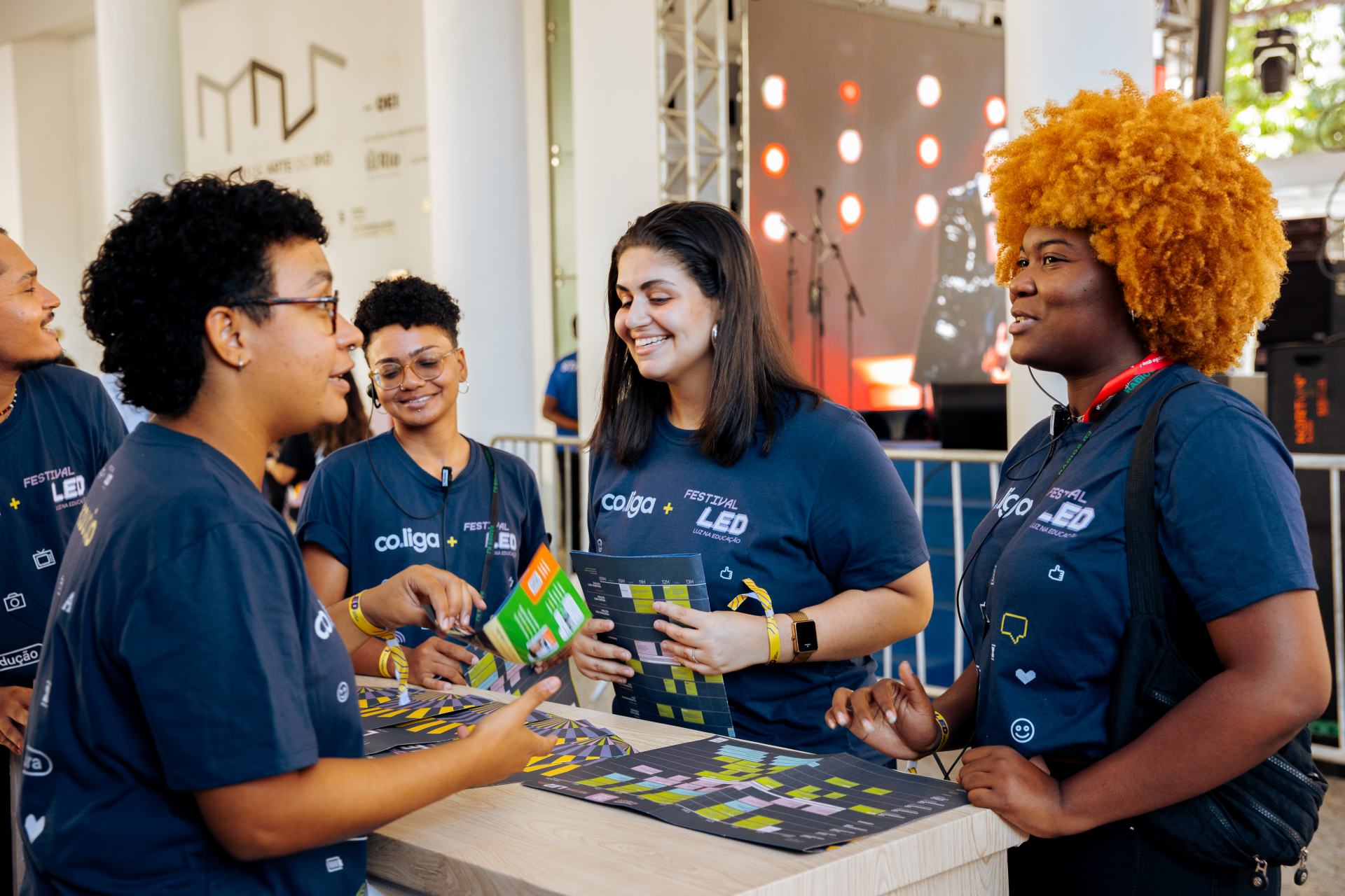 Cinco jovens, três mulheres e dois homens, estão reunidos em um balcão durante um evento. Todos usam camisetas azuis da co.liga no “Festival LED – Luz na Educação” e parecem animados, sorrindo e conversando. Sobre a mesa, há materiais gráficos coloridos. O ambiente tem um palco ao fundo, com luzes vermelhas circulares acesas.