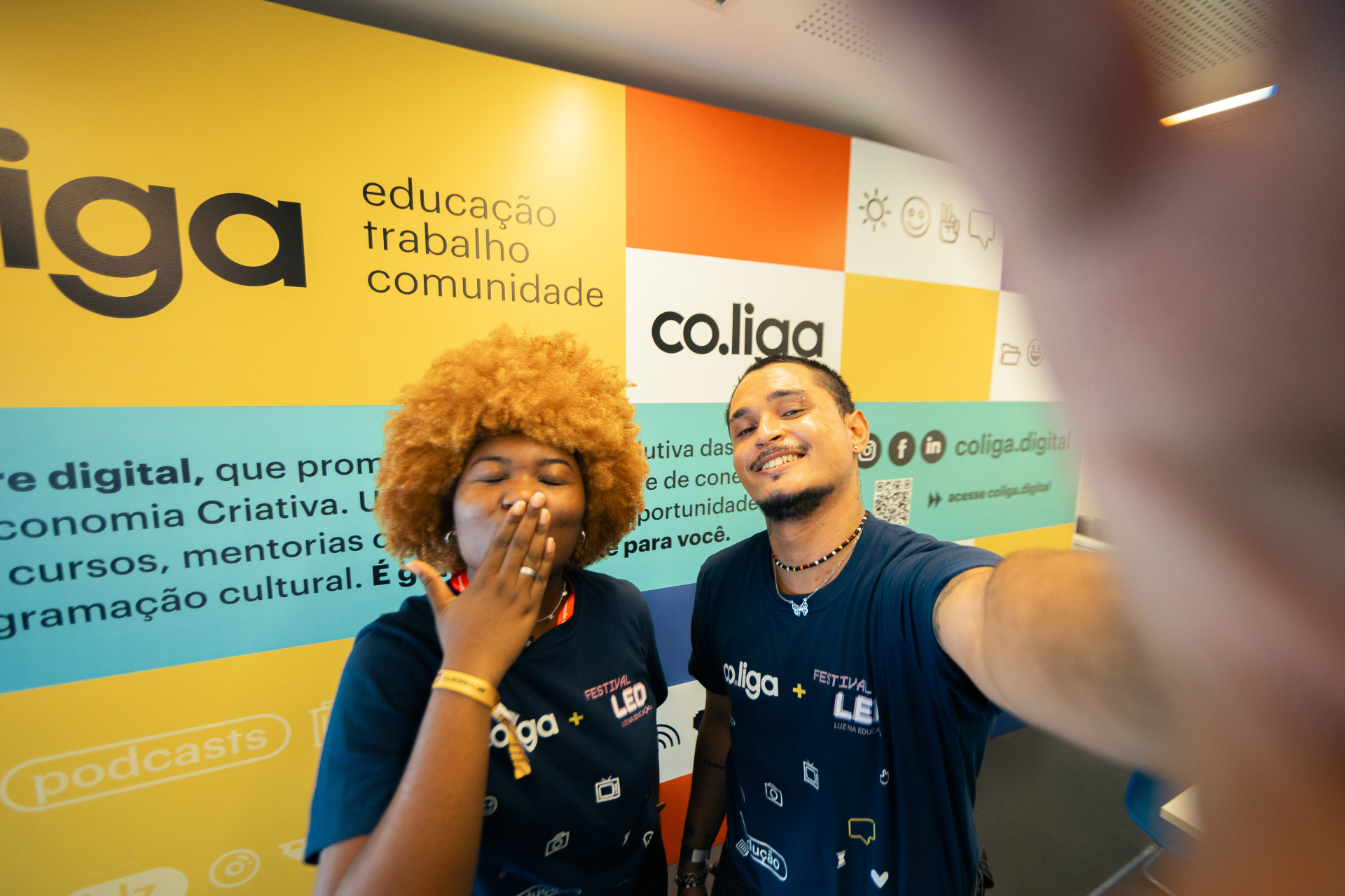 Dois jovens posam sorridentes em frente a uma parede colorida com a identidade visual da plataforma co.liga. À esquerda, uma jovem de cabelos crespos ruivos usa camiseta azul-marinho com o logo da co.liga e envia um beijo com a mão. À direita, um jovem de barba e cabelo preso em coque segura o celular para tirar uma selfie. Ambos usam camisetas com a inscrição "Festival LED". O fundo contém palavras como "educação", "trabalho", "comunidade" e ícones gráficos.