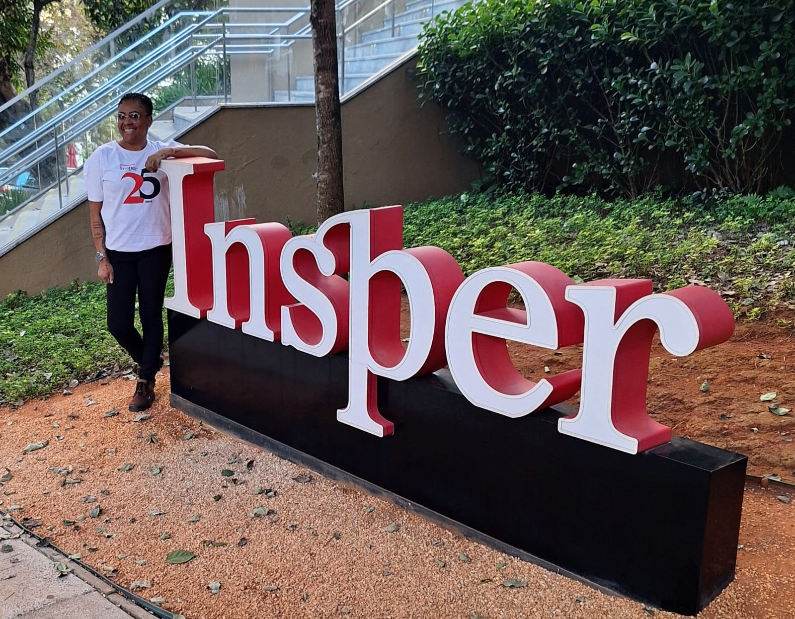 Uma mulher sorridente, com cabelo curto, camisa branca com a estampa de 25 anos e calça preta, posa ao lado de um letreiro grande. O letreiro forma a palavra "Insper" em letras maiúsculas, com as letras "I" e "r" em branco, e as demais em vermelho. O letreiro está sobre uma base preta em um chão de pedriscos.