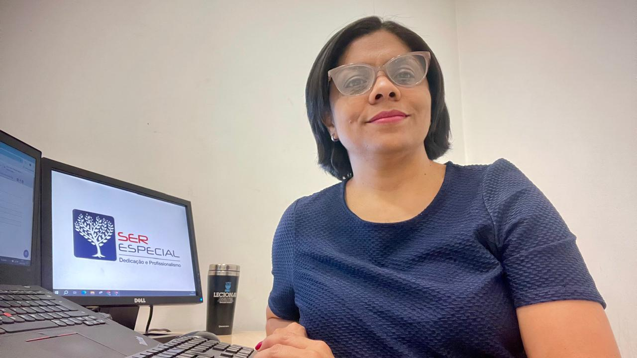 Uma mulher de cabelo escuro, na altura do ombro, e óculos de grau, sentada em uma mesa de escritório. Ela usa uma blusa azul-marinho e está olhando diretamente para a câmera, com um sorriso leve. No fundo, há dois monitores de computador ligados, e um copo térmico sobre a mesa.