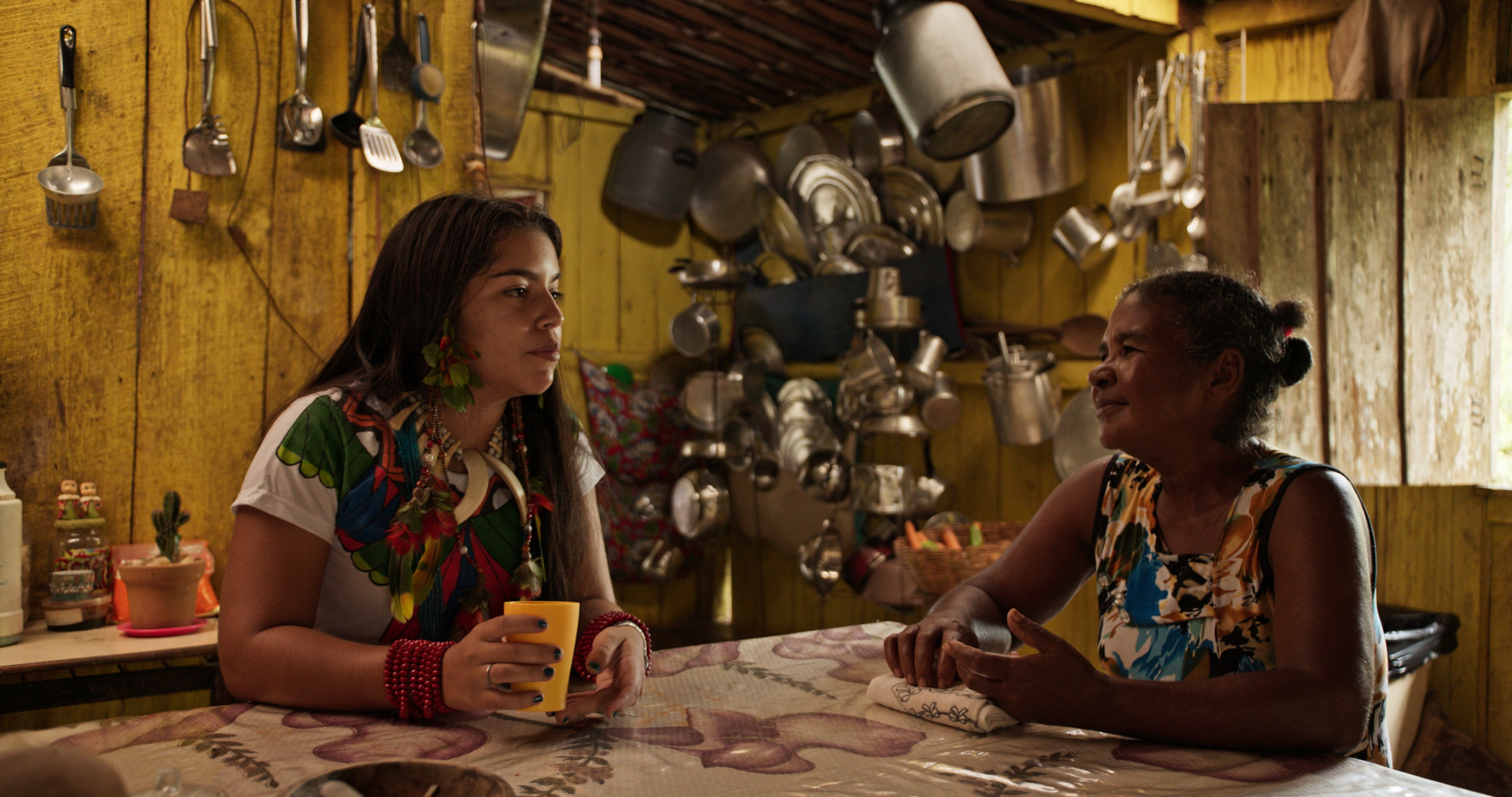 Descrição da imagem: Em uma cozinha simples de madeira pintada de amarelo, duas mulheres estão sentadas frente a frente, conversando à mesa. A mulher à esquerda, mais jovem, tem cabelos longos soltos, usa uma blusa colorida com estampa floral e segura um copo amarelo. Ela também usa brincos grandes e acessórios vermelhos nos pulsos. A mulher à direita, mais velha, tem os cabelos presos e veste uma blusa estampada em tons de azul, branco e laranja; ela apoia os braços sobre a mesa e olha atentamente para a j