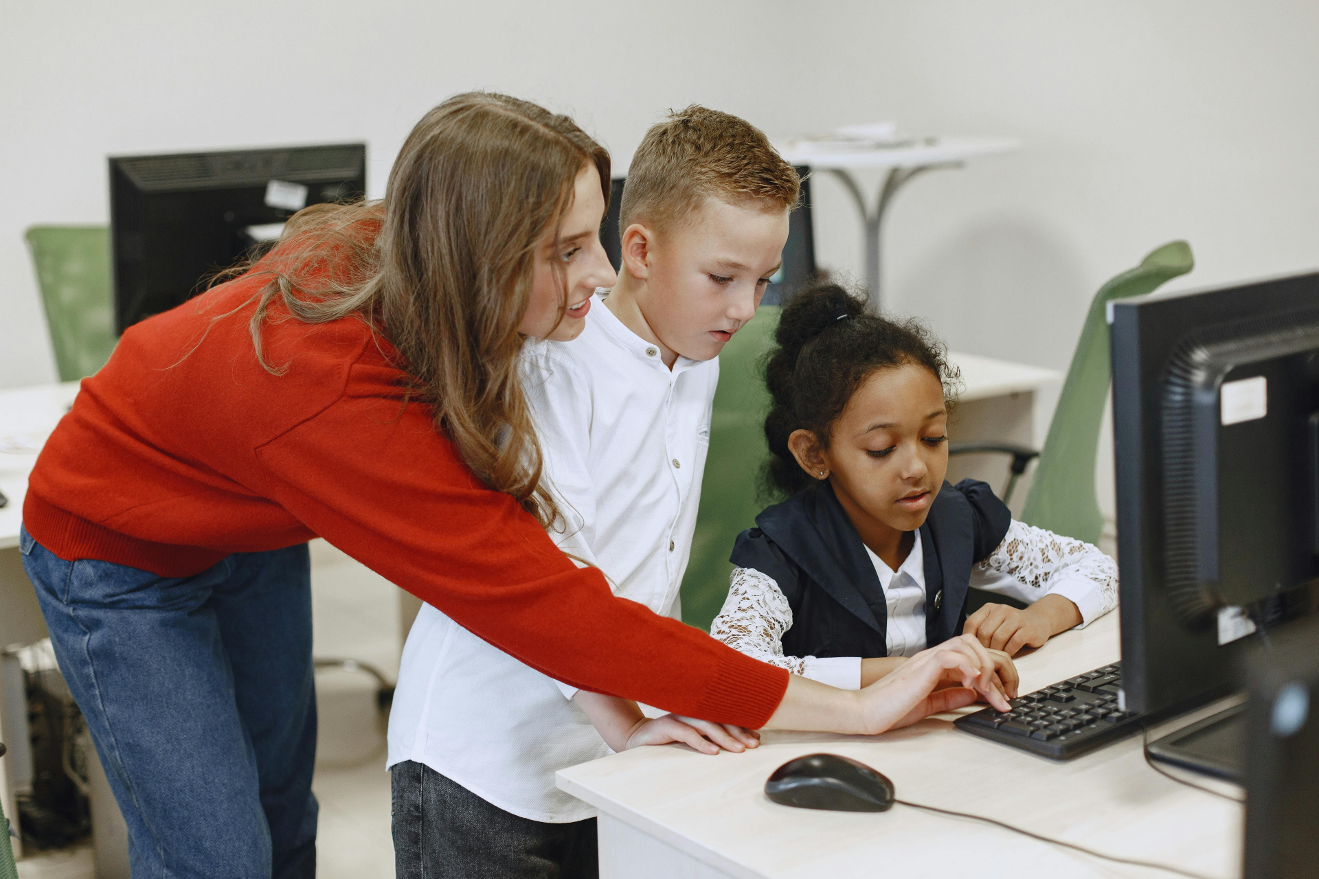 A imagem mostra duas crianças em uma sala de aula usando um computador com o auxílio de uma educadora