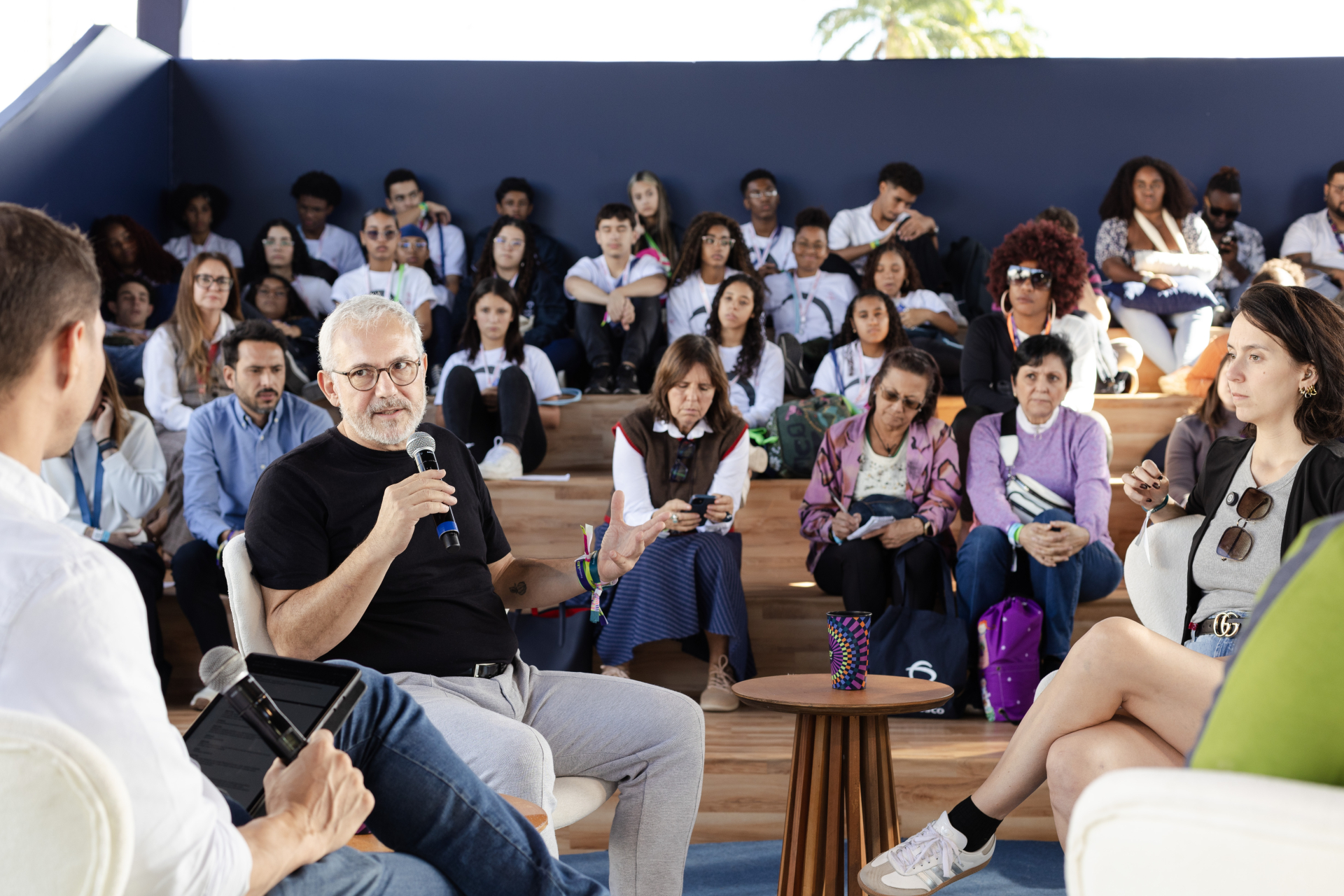 A foto mostra uma roda de conversa em um auditório com arquibancada de madeira. No centro, um homem de barba grisalha e óculos fala ao microfone, gesticulando com a mão, enquanto outras duas pessoas o ouvem — uma mulher jovem à direita e um homem de camisa branca à esquerda, que segura um tablet. Ao fundo, diversas pessoas estão sentadas na arquibancada, entre jovens e adultos, prestando atenção e fazendo anotações. O ambiente é iluminado pela luz natural e tem atmosfera de troca e aprendizado coletivo