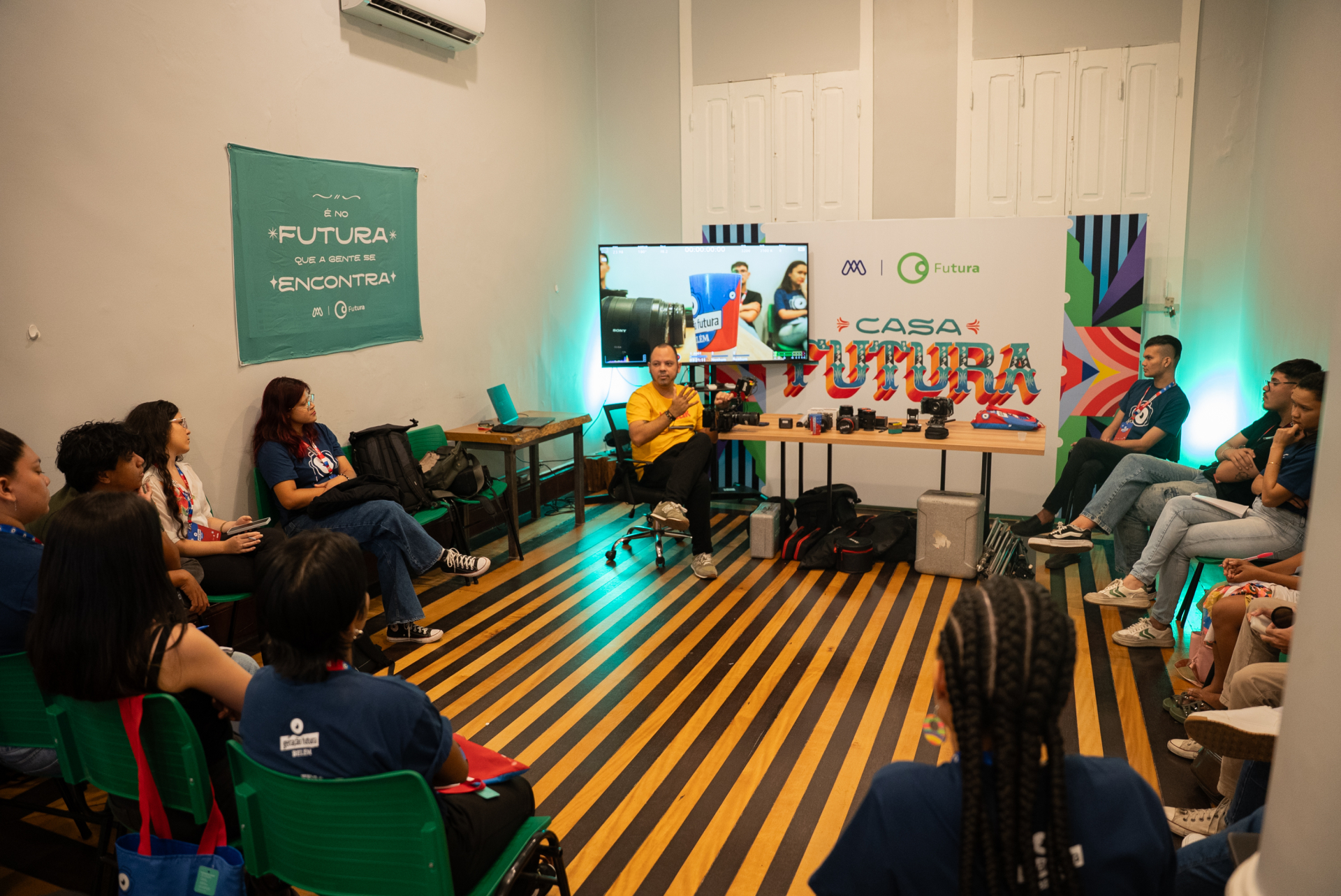 Em uma sala iluminada e colorida, um grupo de jovens participa de uma oficina na Casa Futura. Eles estão sentados em roda, atentos a um instrutor que fala próximo a uma mesa com câmeras e equipamentos audiovisuais. No fundo, o painel exibe o logotipo “Casa Futura” e a frase “É no Futura que a gente se encontra”.