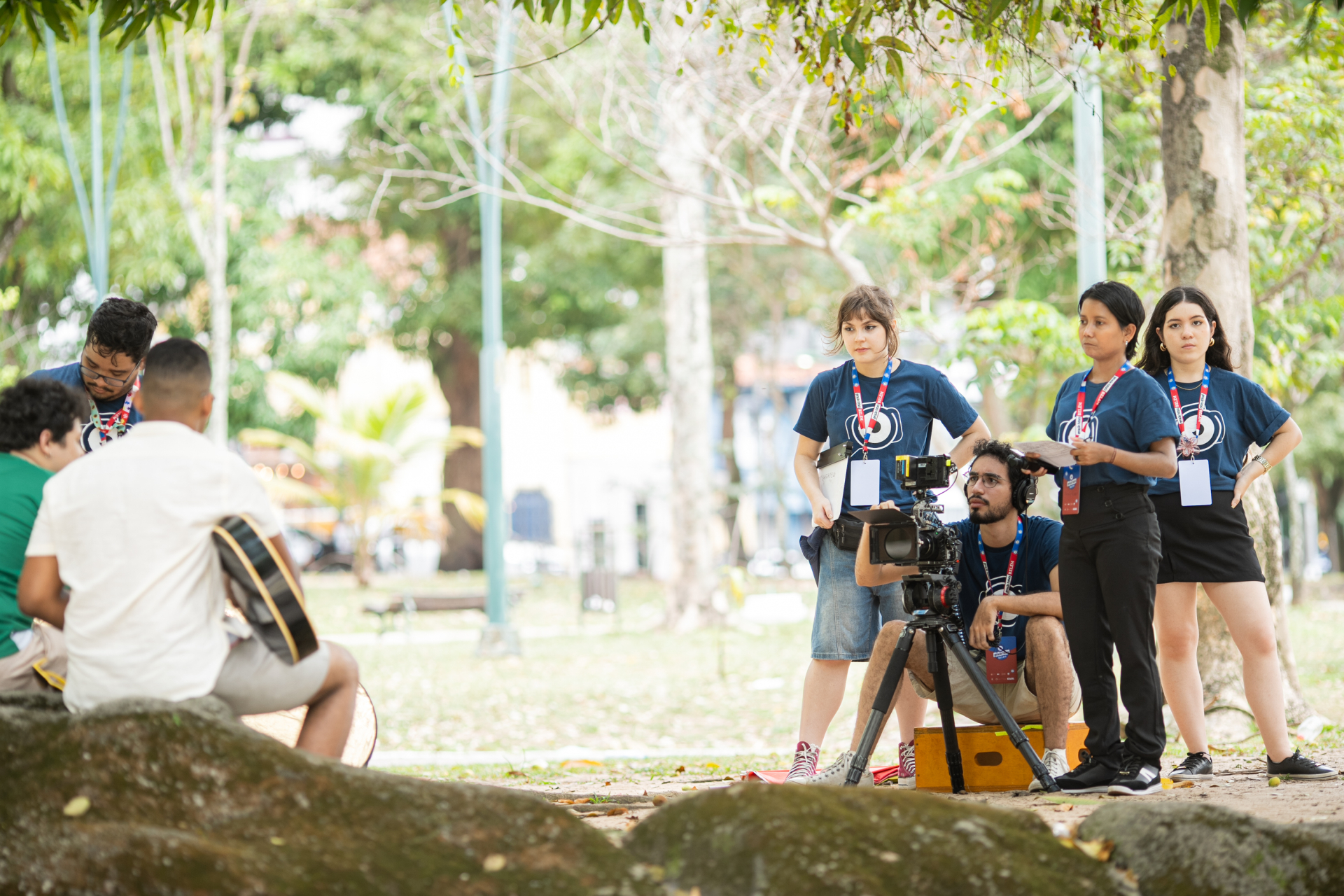 Em um parque arborizado, um grupo de jovens com camisetas azul-marinho está realizando uma gravação audiovisual. No centro da imagem, um rapaz opera uma câmera profissional montada em tripé, enquanto três mulheres e um homem o acompanham com pranchetas e crachás pendurados no pescoço. Ao fundo, dois homens sentados em um tronco de árvore tocam violão, participando da filmagem. A cena transmite concentração e colaboração entre os participantes, em um ambiente ao ar livre e iluminado.