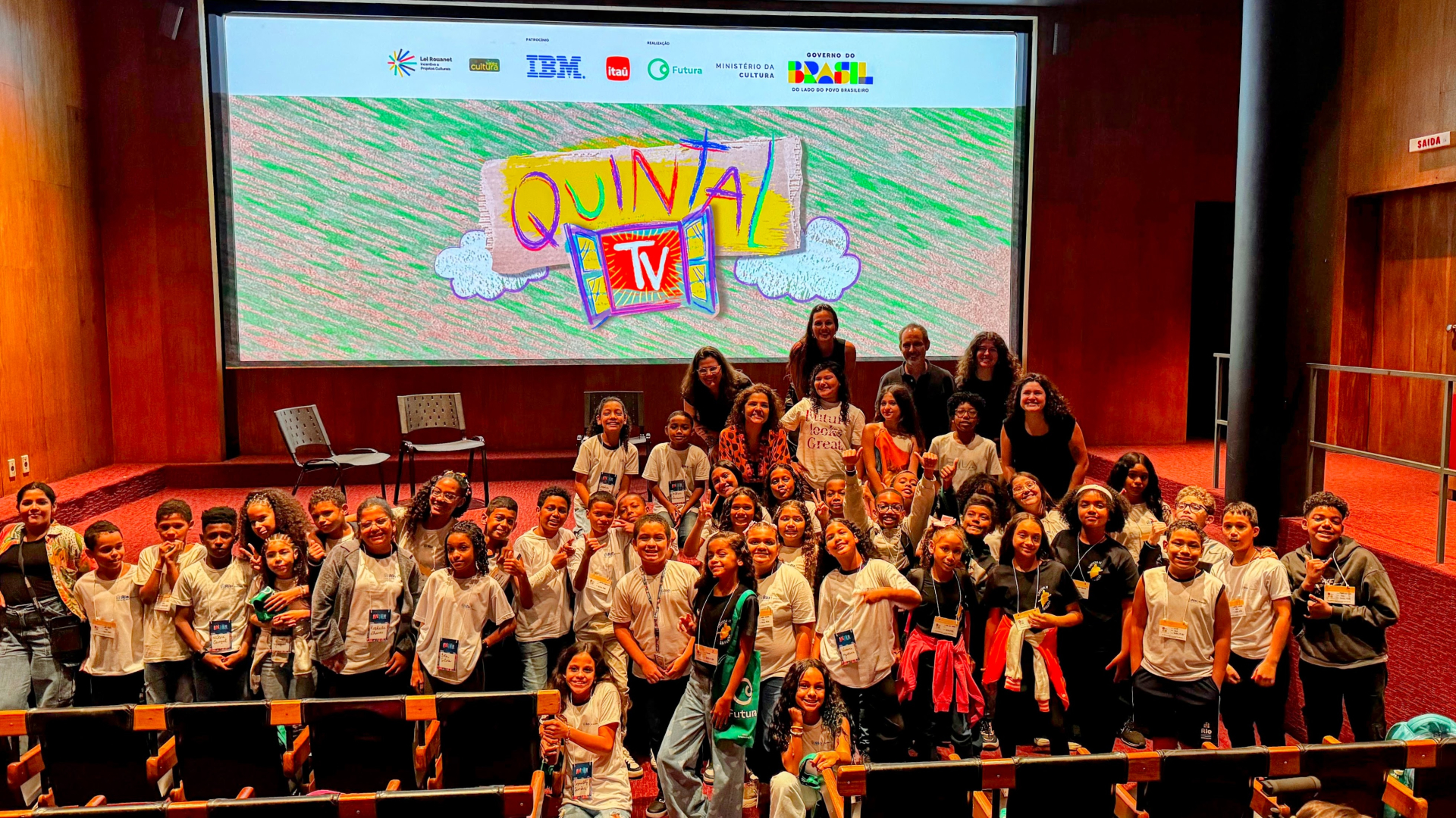 Um grande grupo de cerca de 40 crianças posa para foto diante do mesmo telão com o logotipo Quintal TV. Elas estão sorrindo e posam animadas ao lado de adultos, possivelmente educadores e organizadores do evento. A maioria veste camisetas claras com crachás de identificação. O cenário é o mesmo auditório da primeira imagem, com paredes de madeira e piso avermelhado.