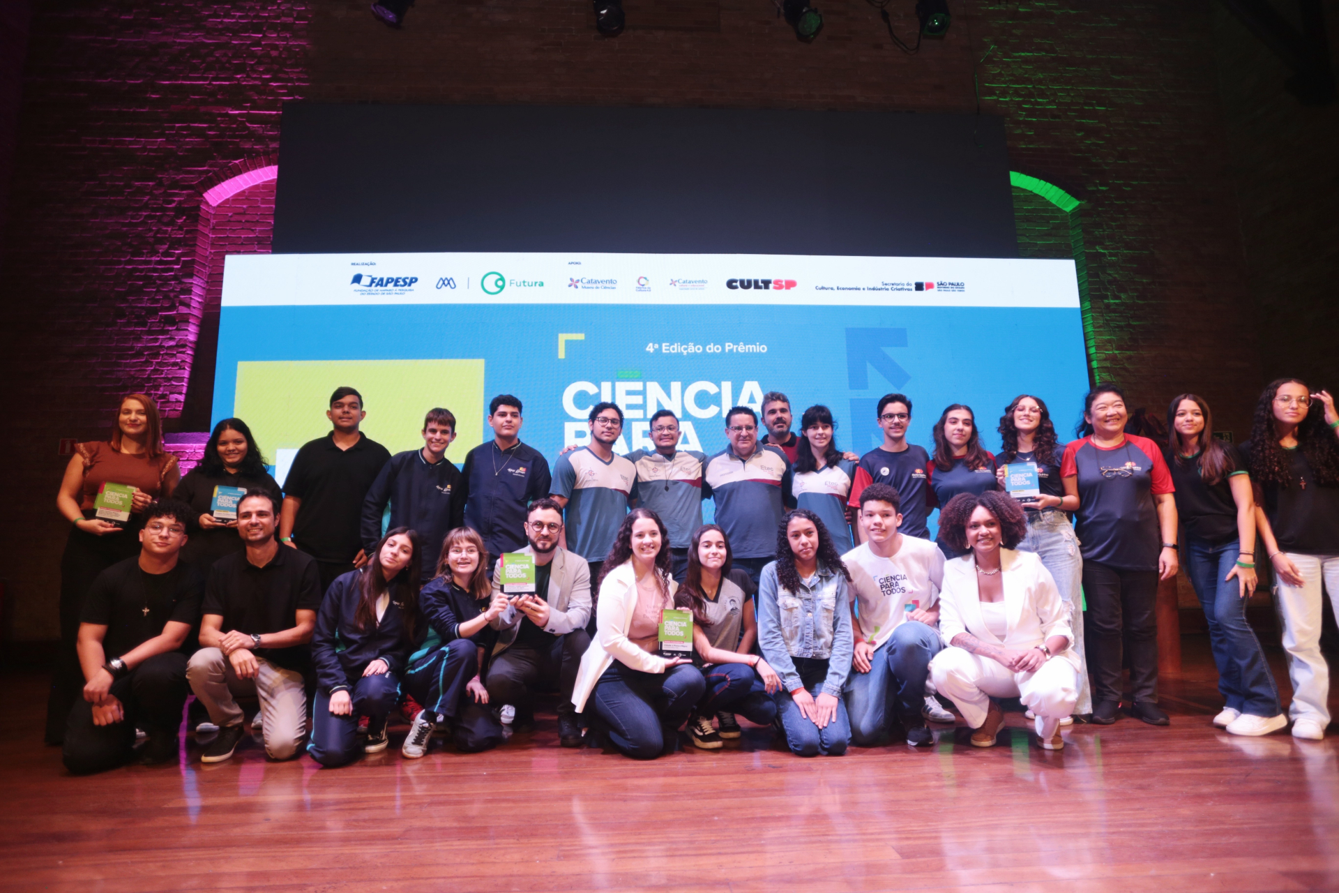Um grande grupo, formado por jovens estudantes, professores e convidados, está reunido sobre um palco de madeira. Ao fundo, um telão azul mostra a identidade visual da 4ª edição do Prêmio Ciência Para Todos. Algumas pessoas seguram placas ou troféus nas mãos. O grupo está organizado em duas fileiras: parte em pé e parte agachada ou sentada. O clima é festivo, e todos olham para a câmera, muitos sorrindo. O ambiente tem paredes de tijolos à vista, com iluminação roxa e azul.