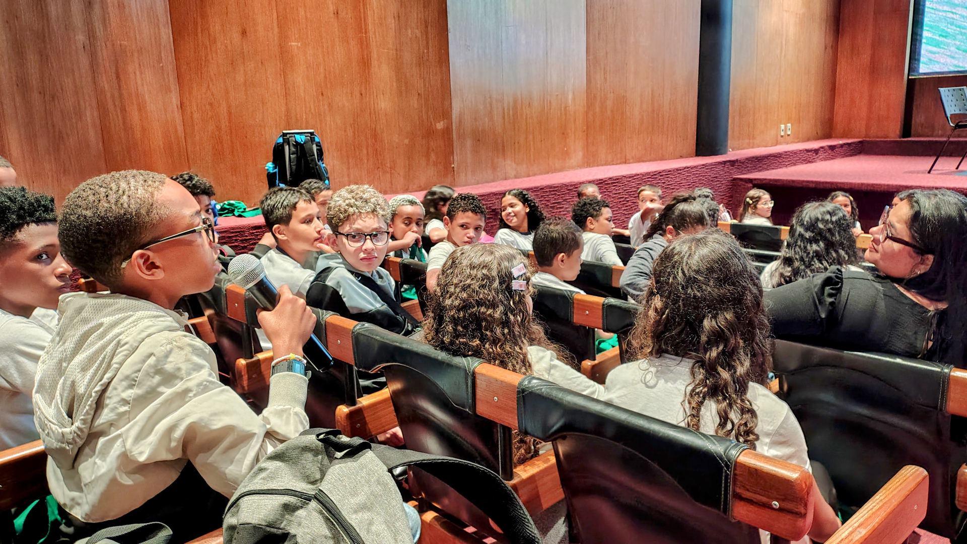 Crianças estão sentadas em cadeiras de um auditório, voltadas para o palco. Um menino em destaque, segurando um microfone, faz uma pergunta ou comentário. Outras crianças ao redor o observam com atenção. Ao fundo, uma mulher parece ouvir atentamente. As paredes do local são de madeira e o ambiente tem iluminação suave.