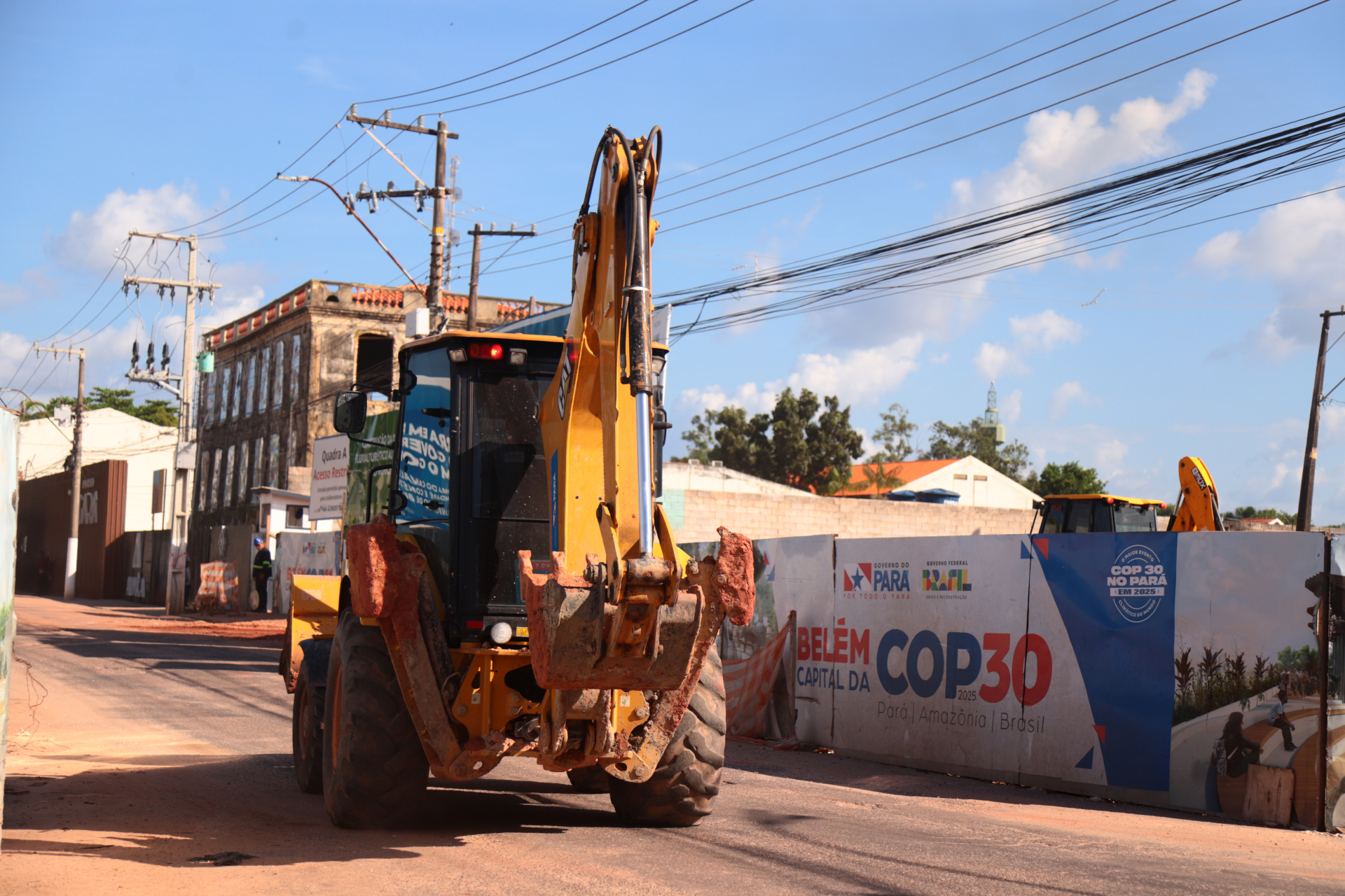 Uma retroescavadeira amarela avança por uma rua em obras em Belém. À direita, um tapume traz os dizeres “Belém capital da COP30 – Pará, Amazônia, Brasil”, com logotipos do Governo do Pará. O céu está azul e o cenário mistura construções antigas e novas, simbolizando transformação urbana.