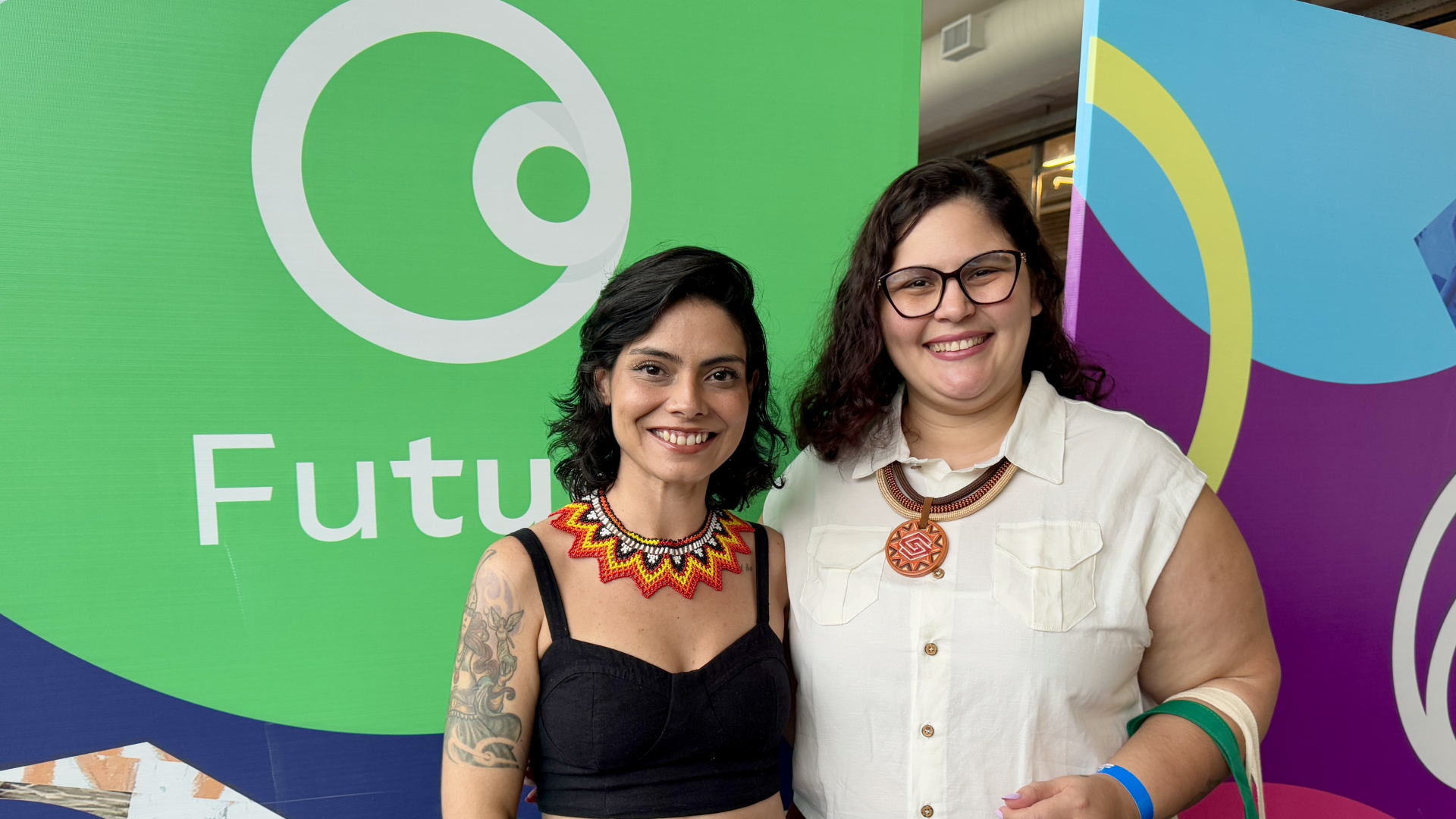 Duas mulheres sorriem lado a lado diante de um painel colorido com o logotipo da Casa Futura. Ambas usam colares artesanais de inspiração indígena; uma veste blusa preta e a outra, camisa branca sem mangas. O fundo tem cores vivas — verde, roxo e azul — que reforçam o clima alegre e acolhedor do ambiente.
